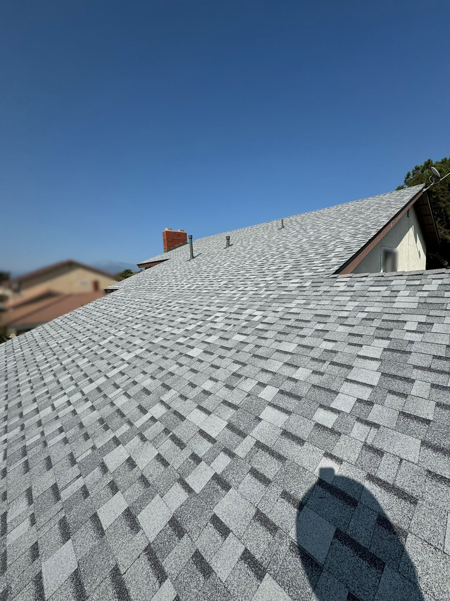 Gray and white asphalt shingles on a rooftop under a clear blue sky. A brick chimney is visible.