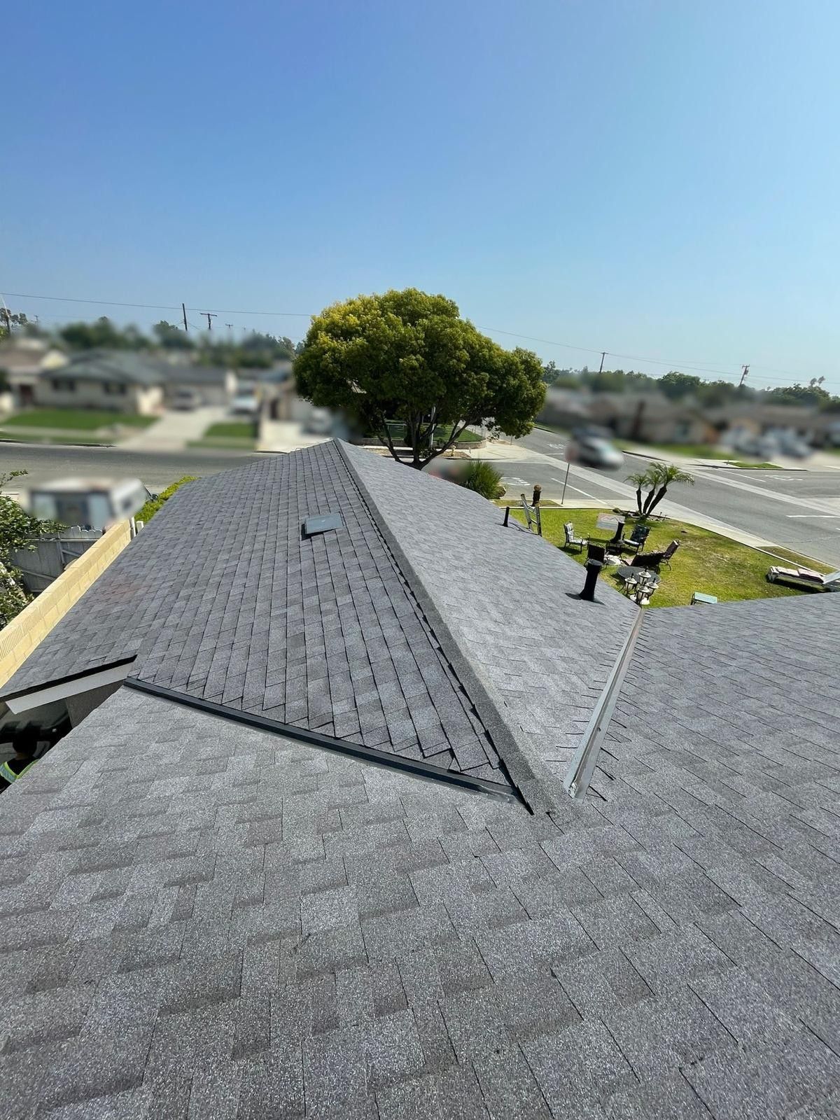 Gray shingle roof with a tree and houses in the background on a sunny day.