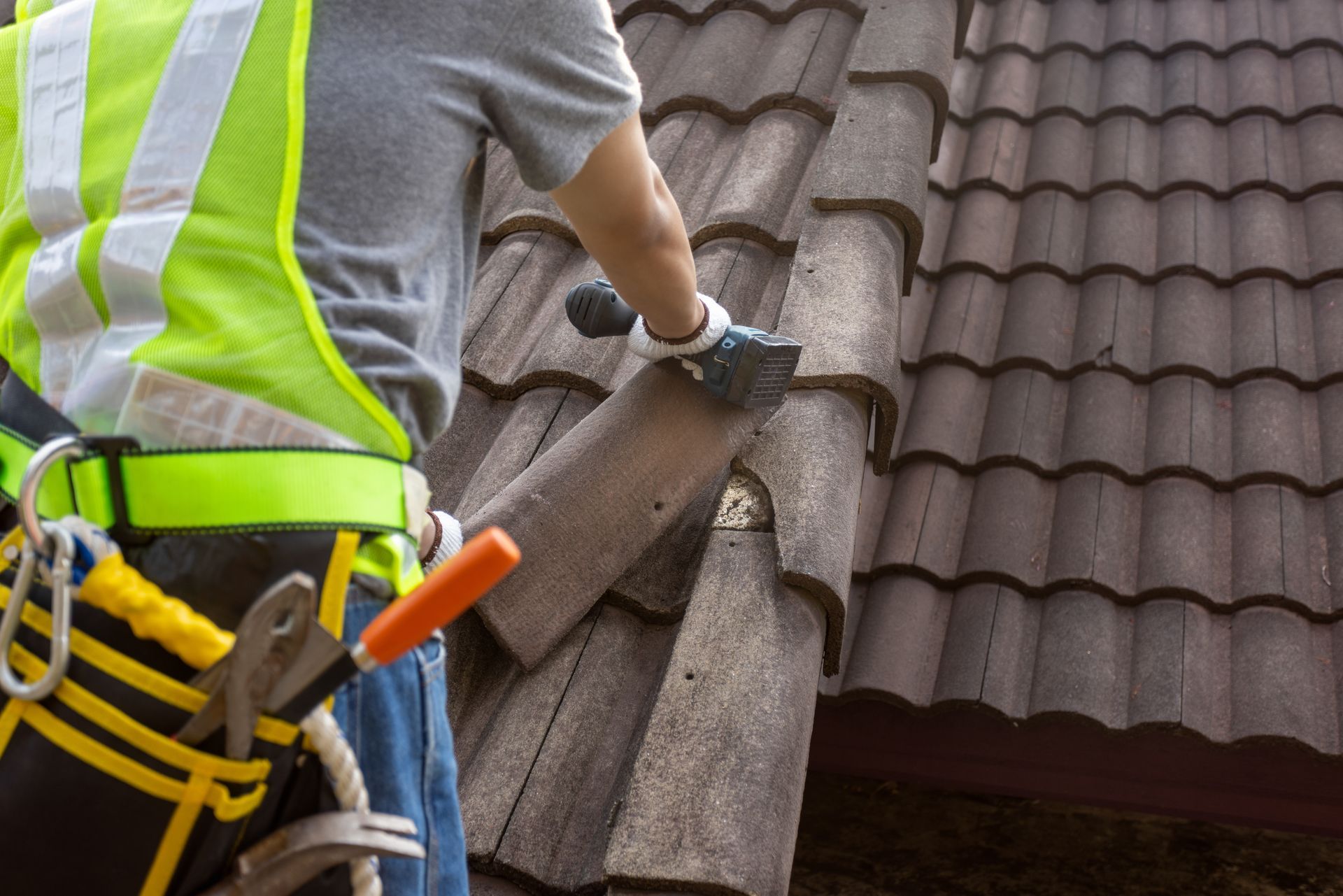 Roofer in safety vest replacing a weathered tile on a roof; safety harness, tool belt visible.