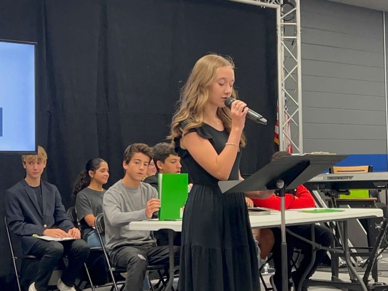 Girl in black dress speaks into a microphone at a podium. Audience sits behind.