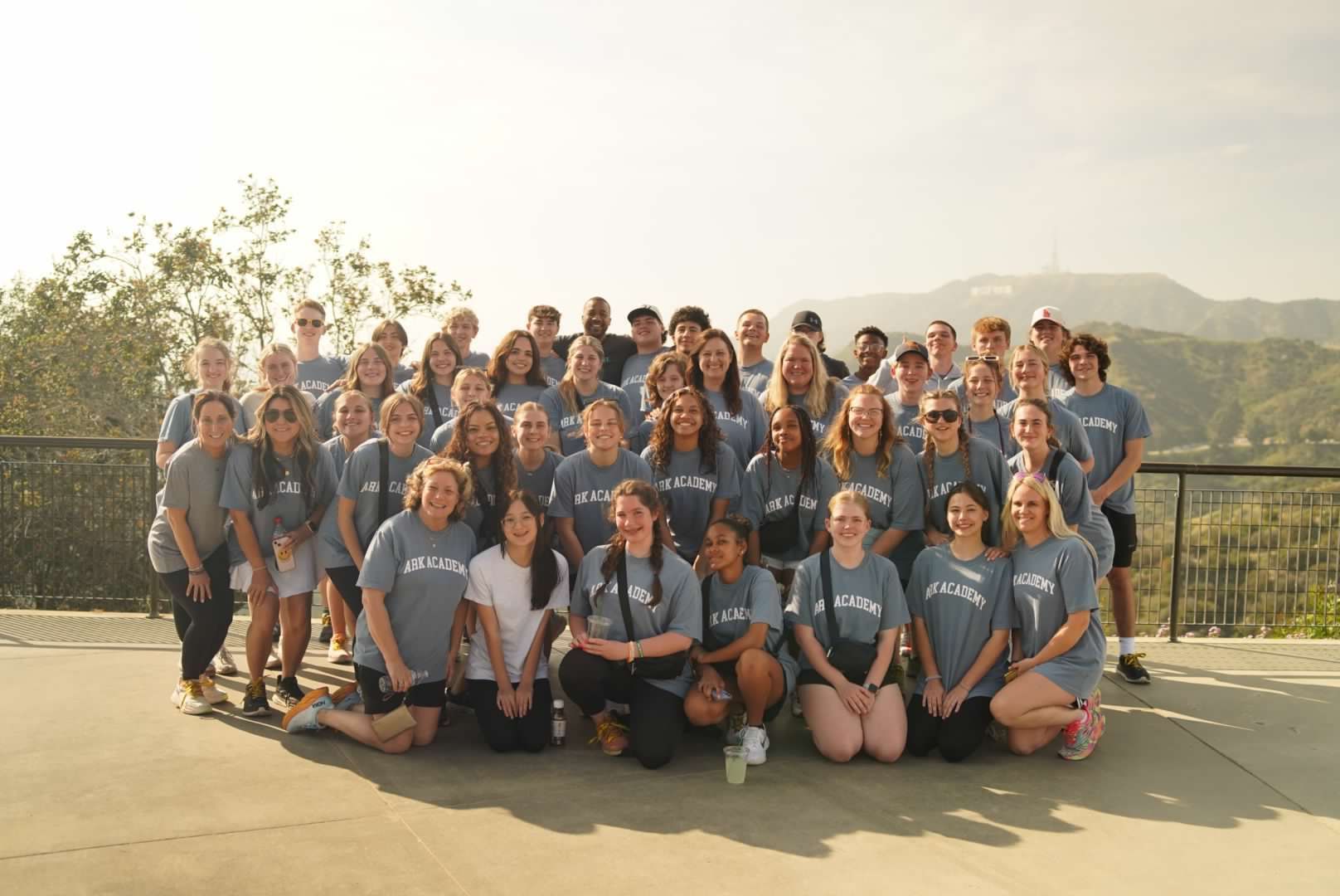 Group of people in gray shirts pose outdoors with Hollywood sign in background.