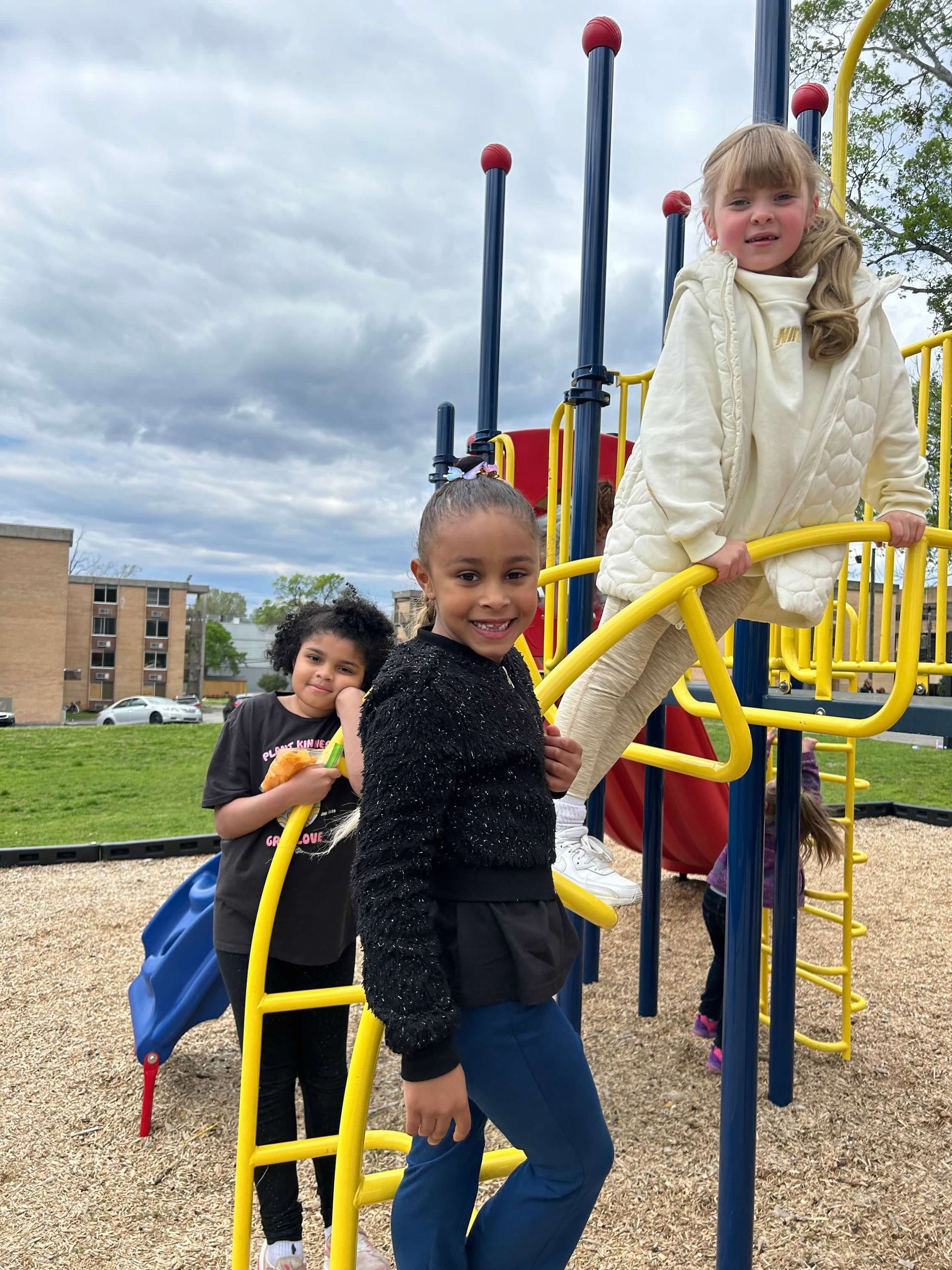 Children playing on a colorful playground. One is smiling, climbing the monkey bars, and other children are nearby.