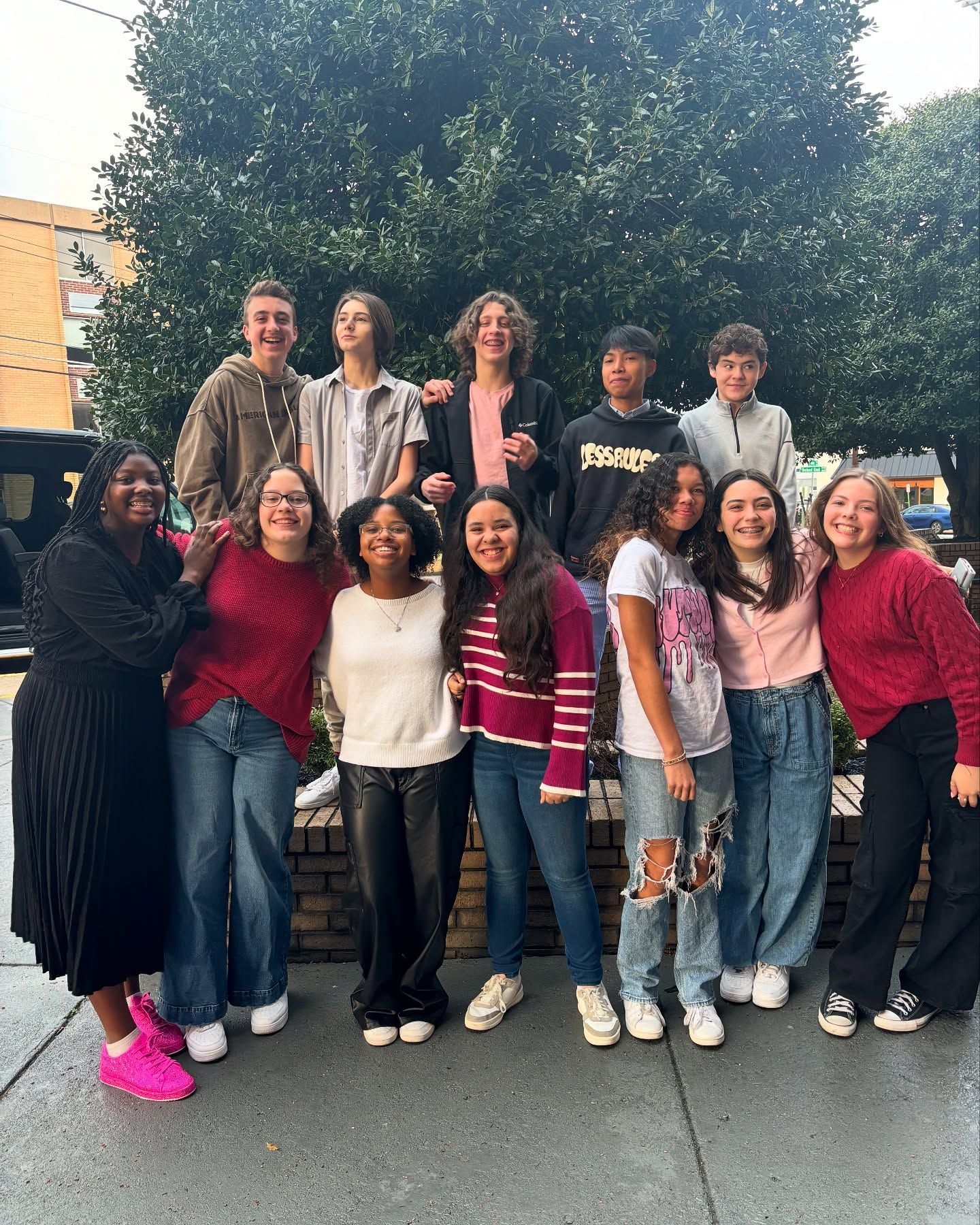 Group of students smiling together outdoors in front of a hedge, some are wearing sweaters and jeans.