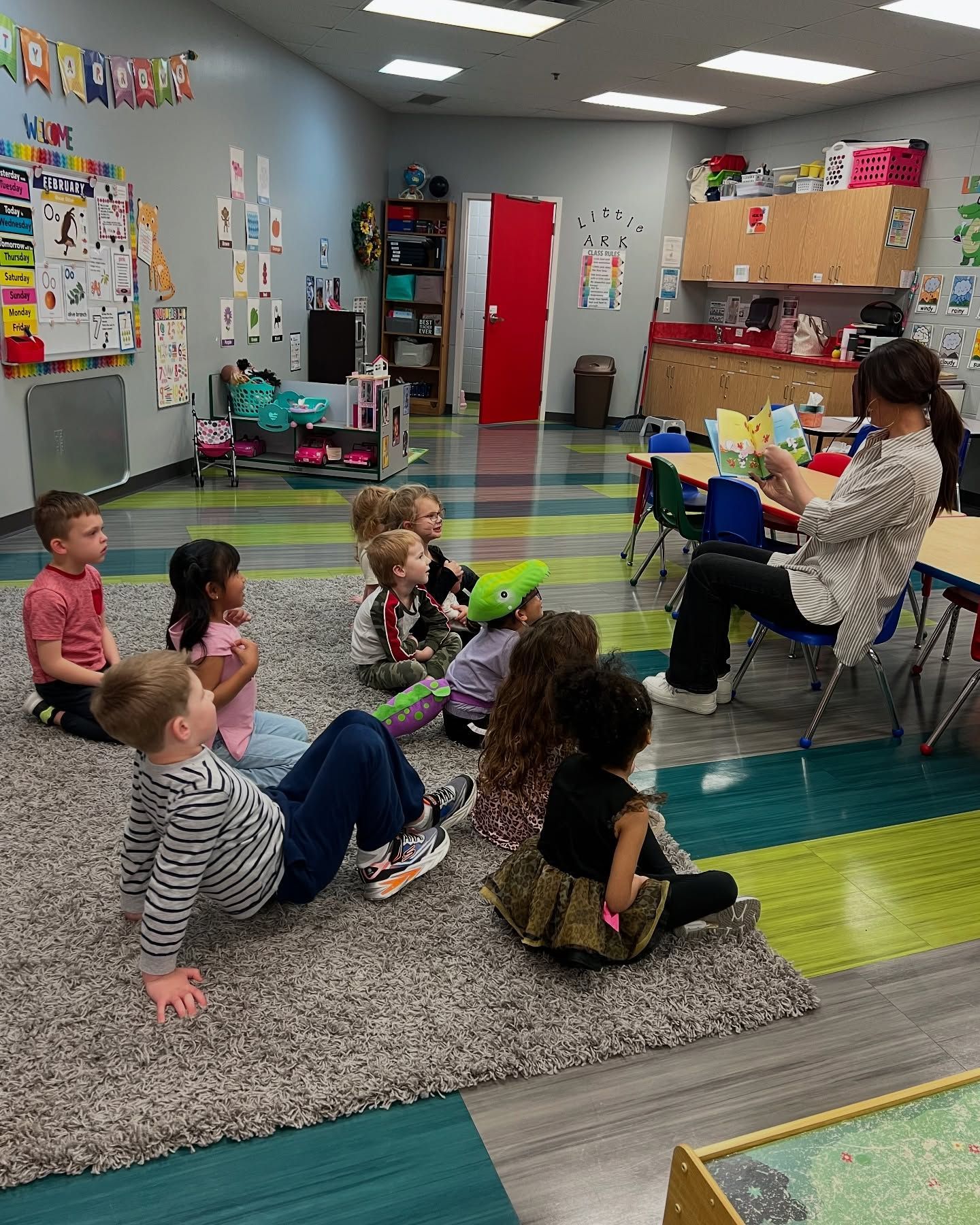 Children sitting on a rug, listening to a person reading a book in a classroom.