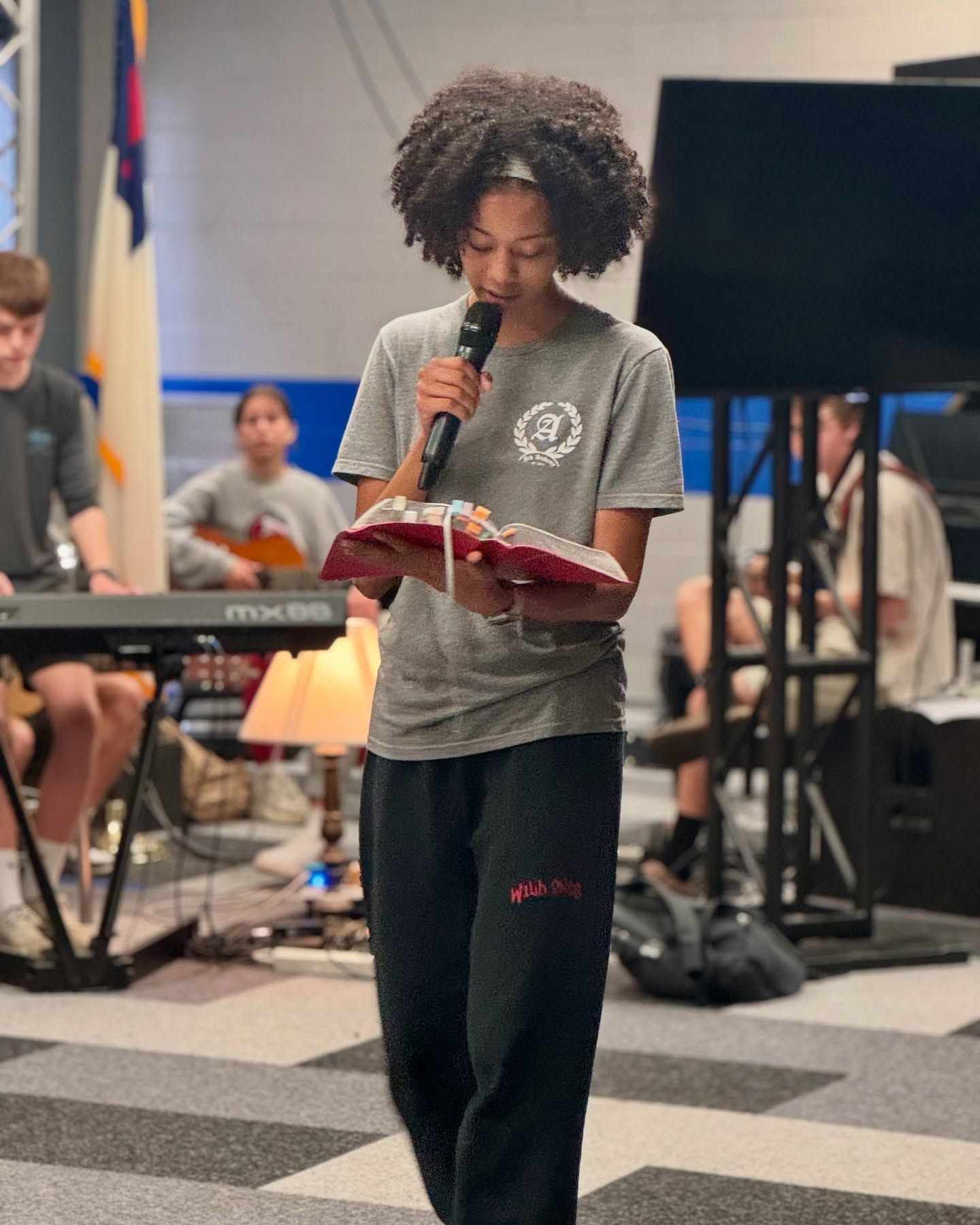 Young person with curly hair speaking into a microphone while reading from a book, possibly a church setting.