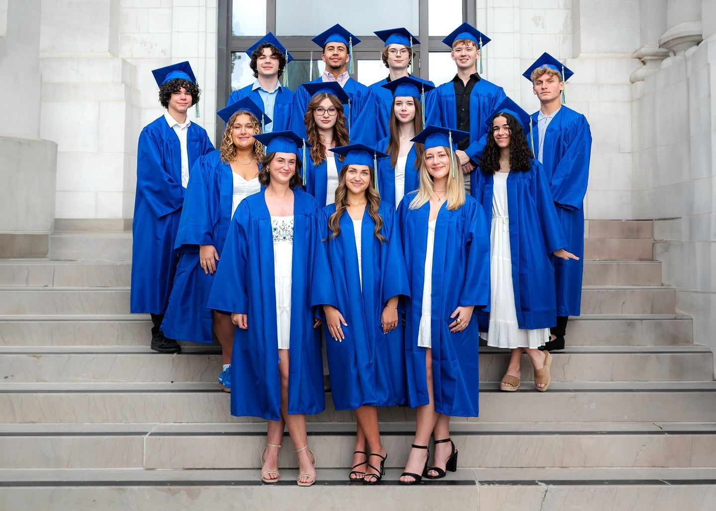 Group of graduates in blue gowns and caps, standing on stone steps of a building.