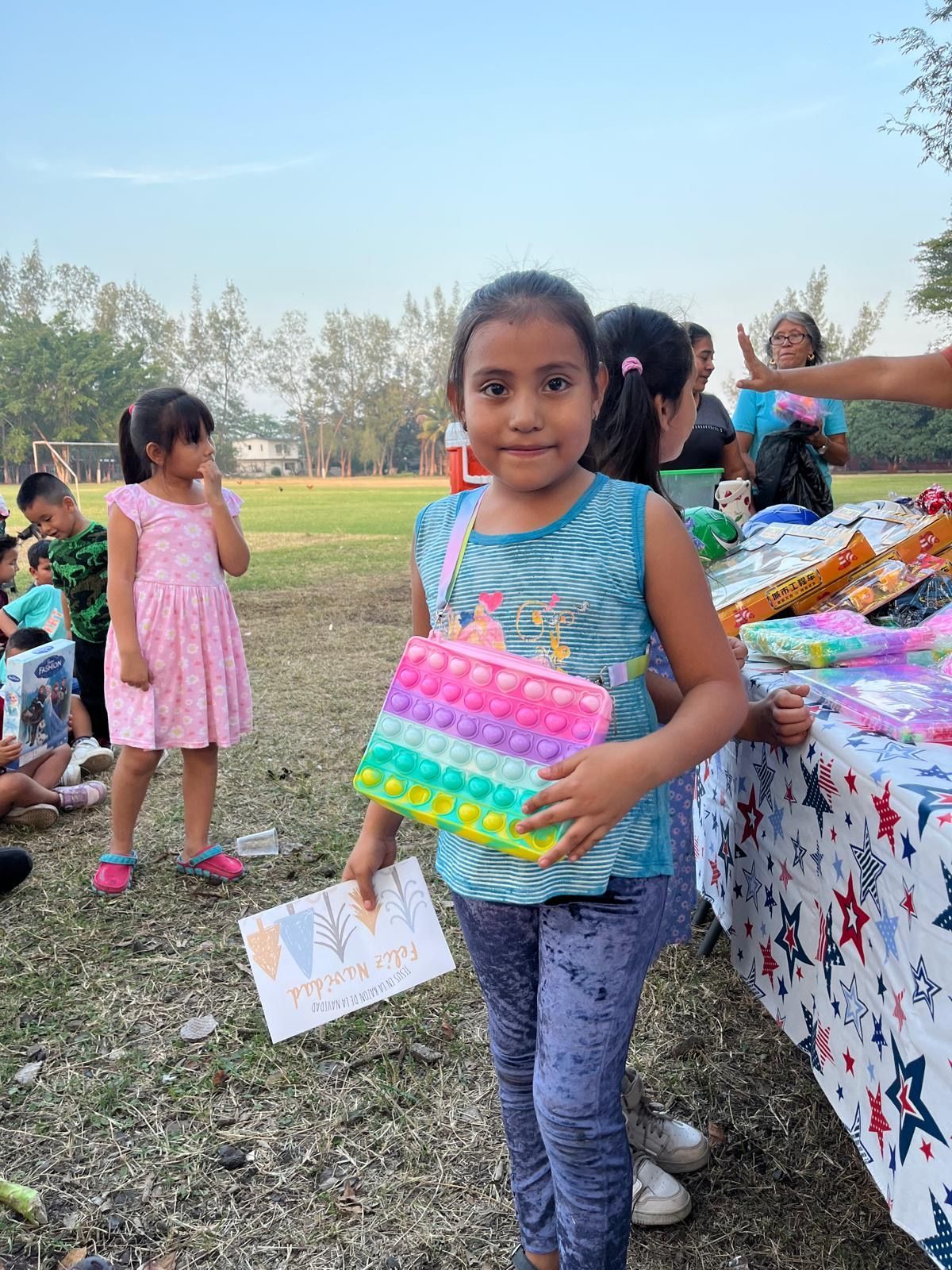 Girl holding a colorful pop-it purse and a certificate outdoors, smiling at the camera.