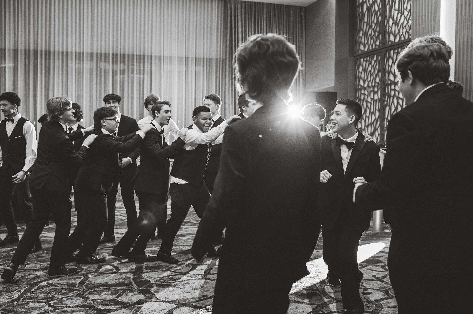 Group of people in suits dancing in a room with patterned carpet and large windows.