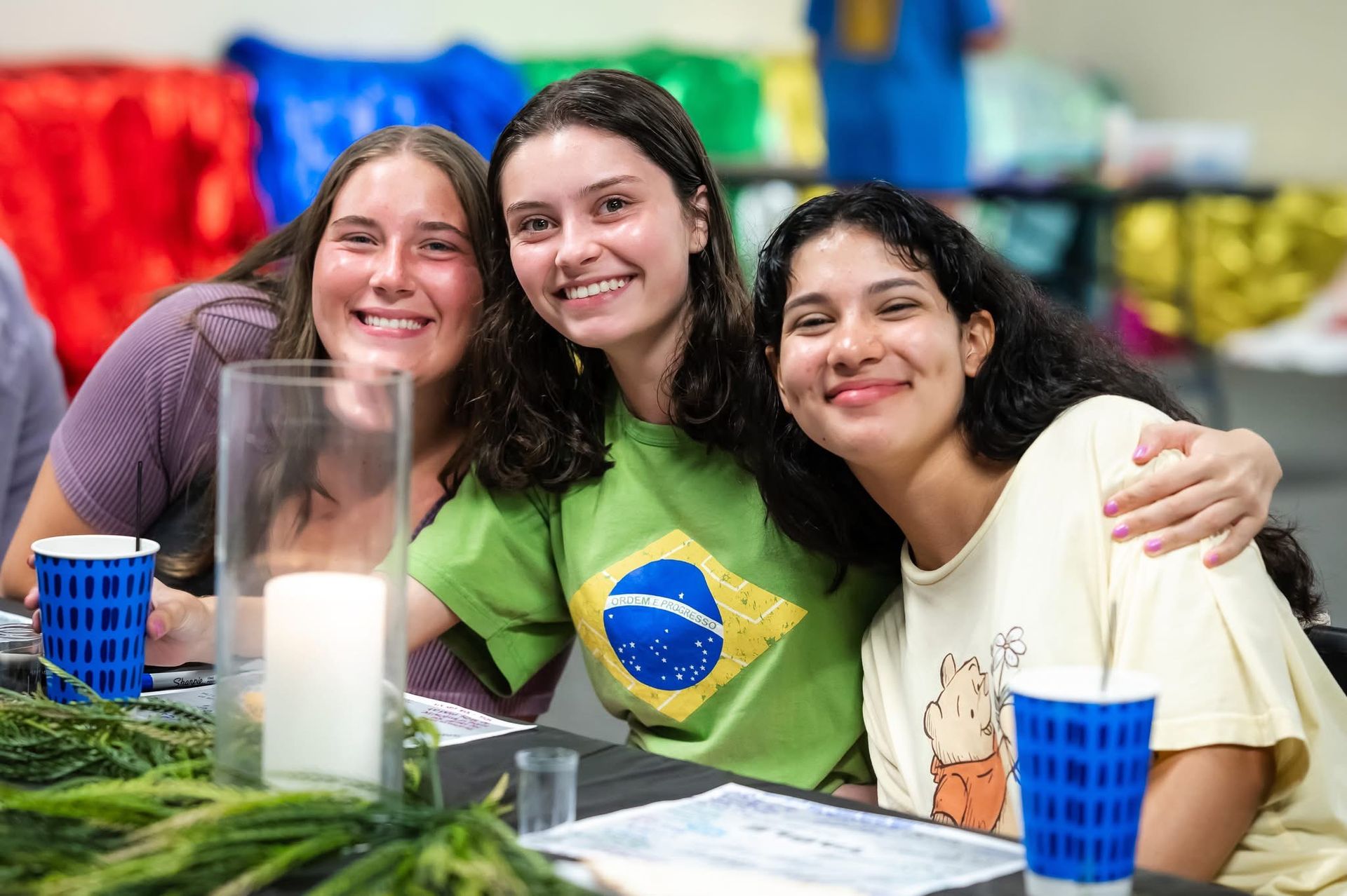Three smiling people at a table; one wears a green shirt with a Brazilian flag.