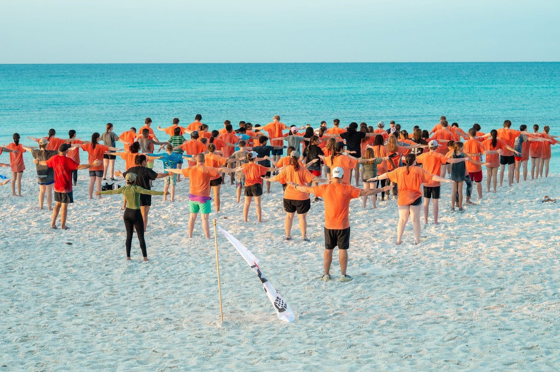 Group of people in orange shirts with arms outstretched on a beach, facing the ocean.