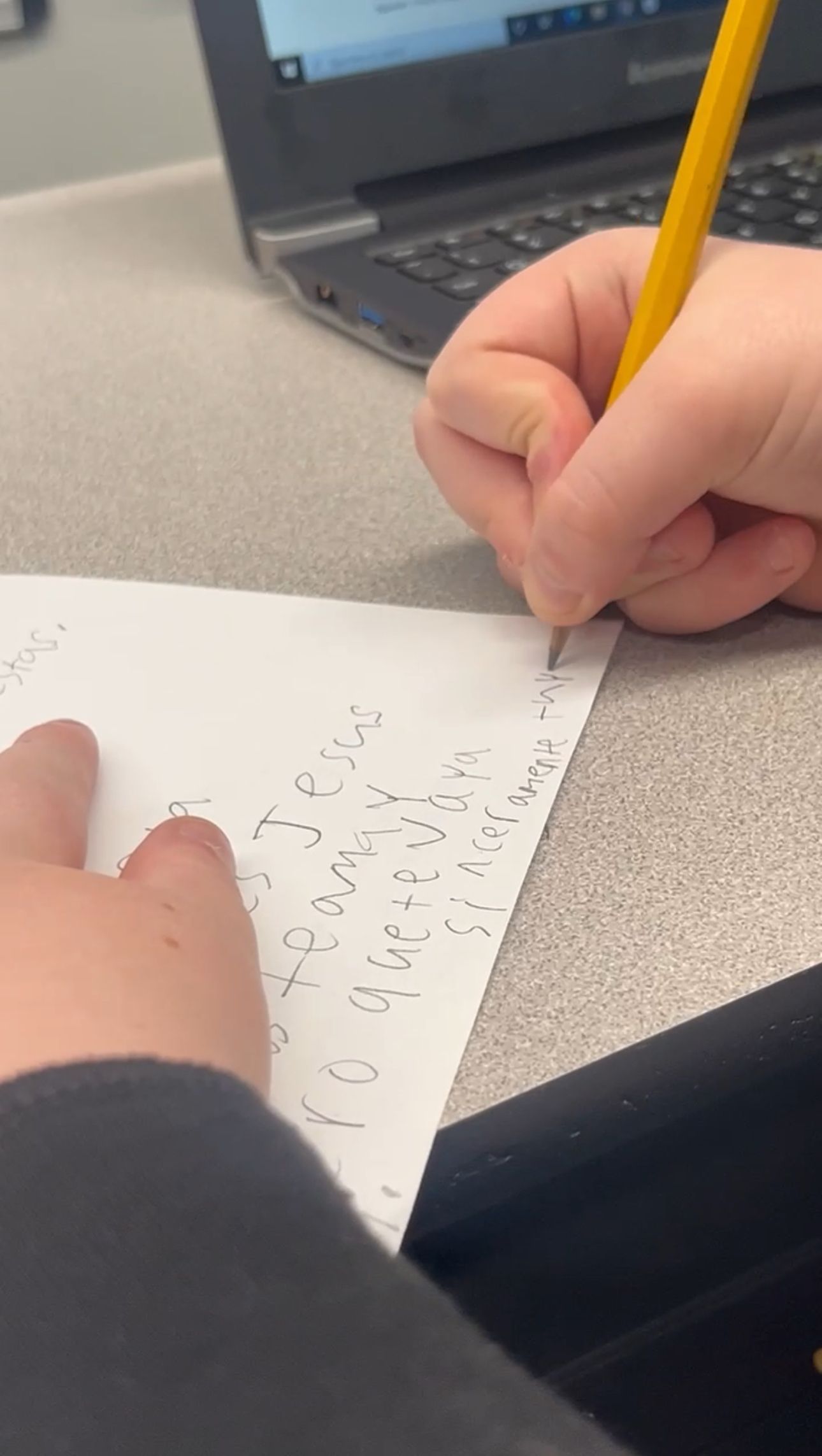 Person writing on paper with a pencil, laptop in the background.