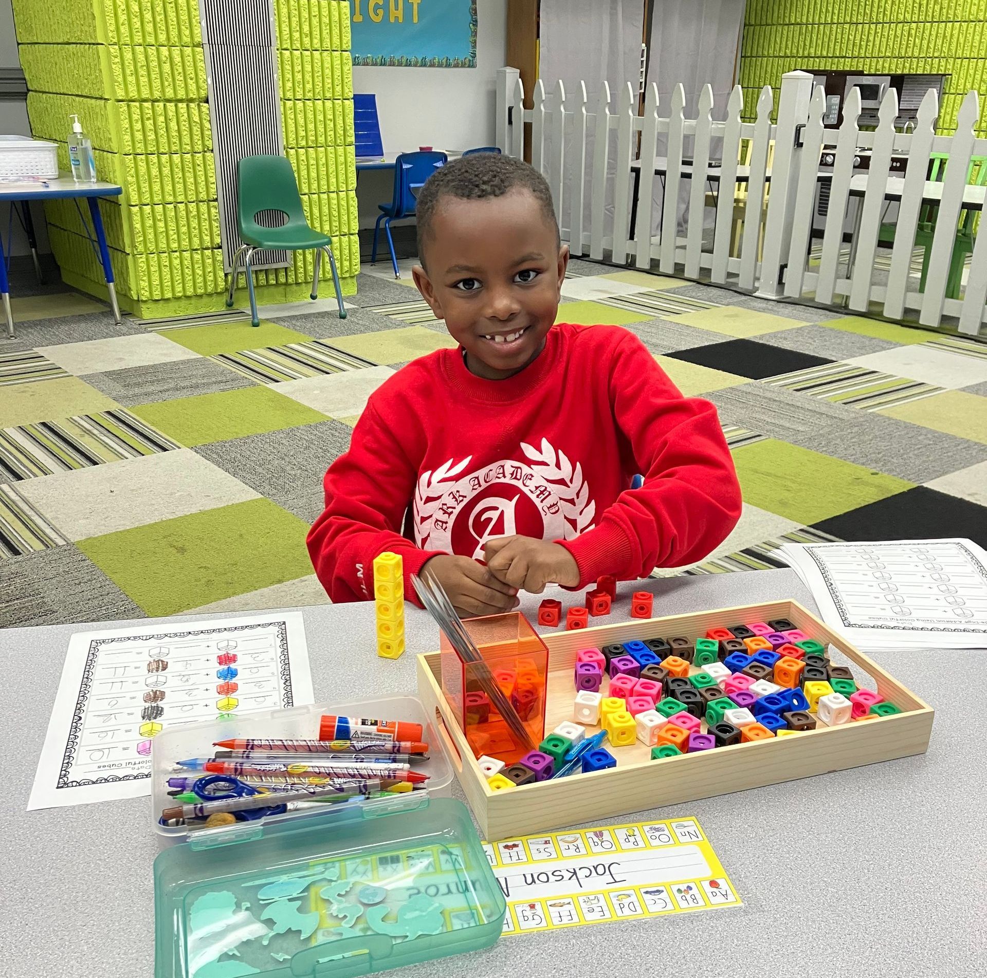 Young child seated at a desk, smiling while playing with colorful blocks in a classroom.