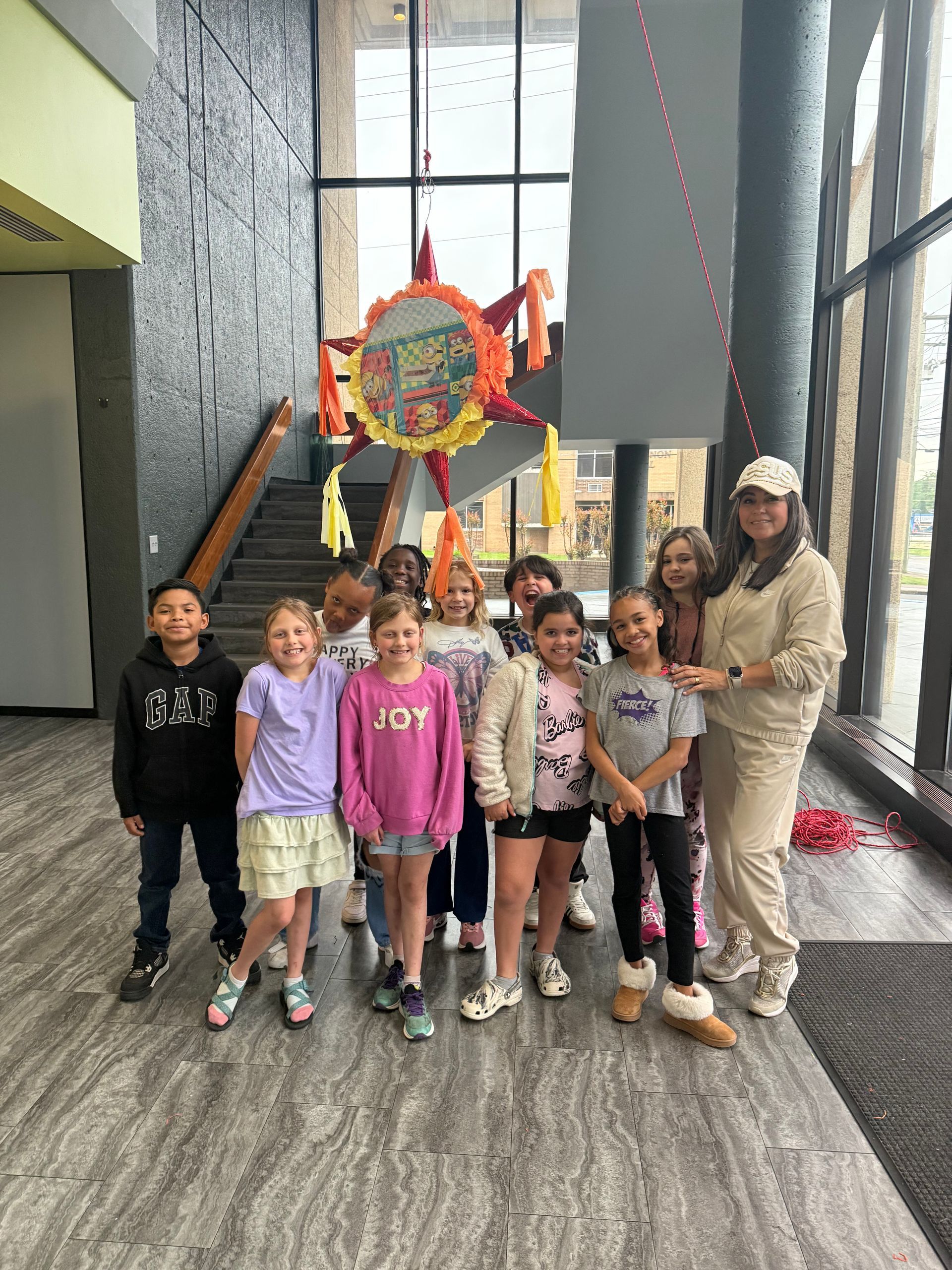 Group of children with a woman by a piñata in a building. Children are smiling.