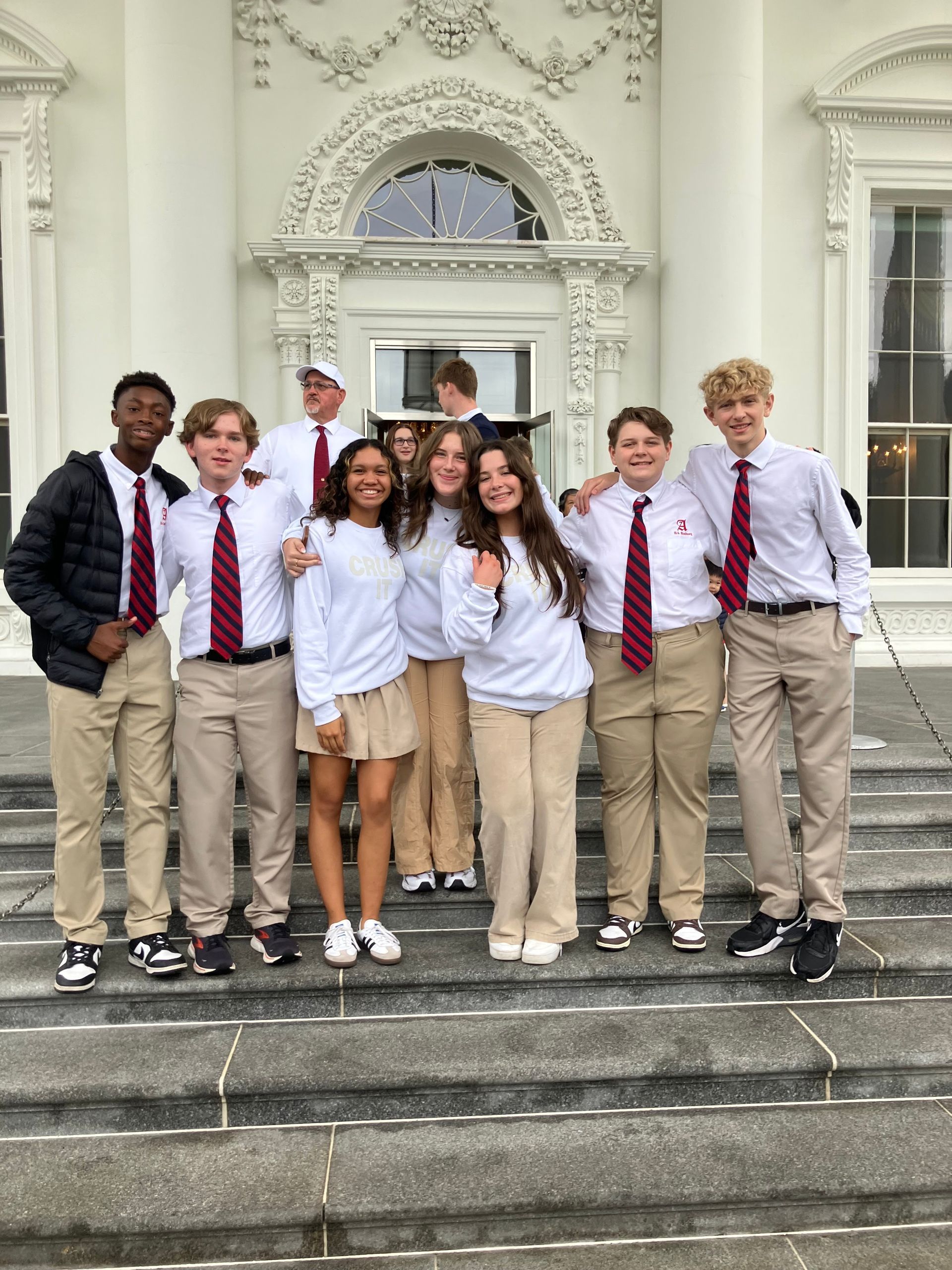 Group of students in uniform pose on steps in front of a white building with ornate details.