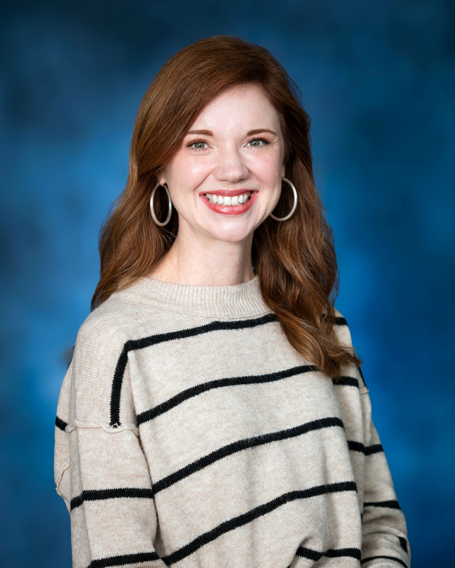 Woman with red hair smiling, wearing a striped sweater and hoop earrings, against a blue background.