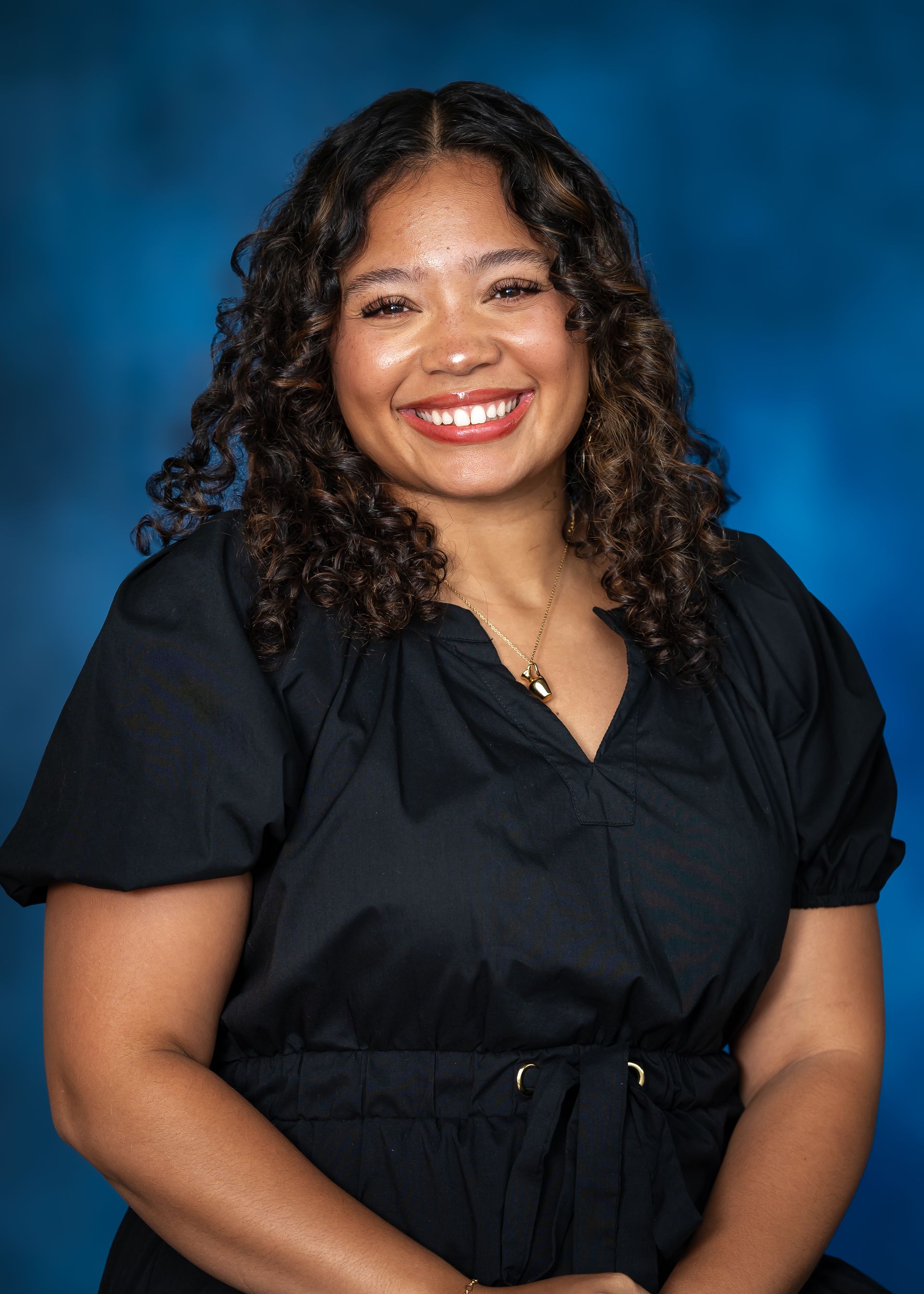 Smiling person with dark curly hair in a black top against a blue backdrop.