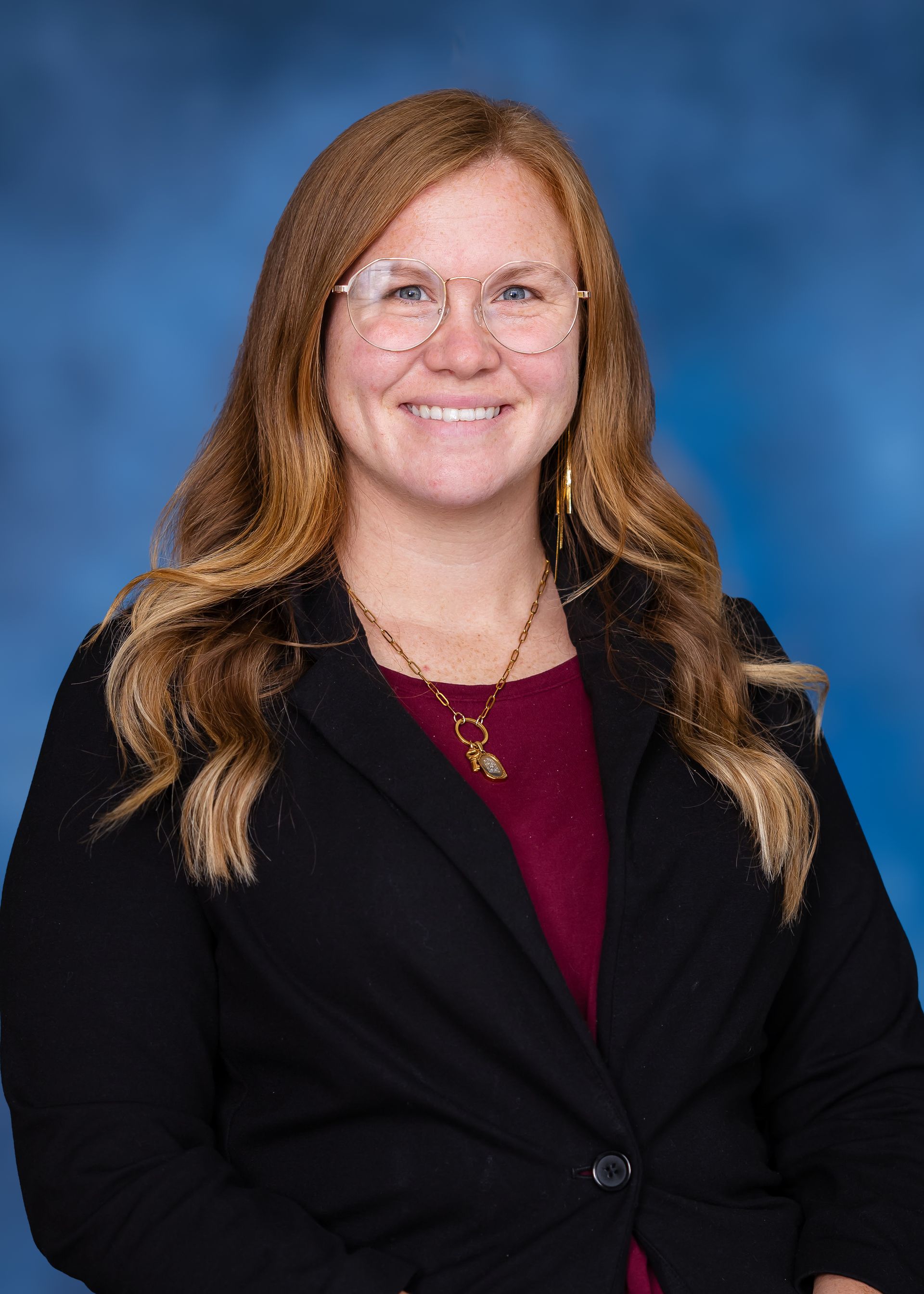 Woman with glasses, long auburn hair, wearing a black blazer and maroon top, smiling against a blue backdrop.