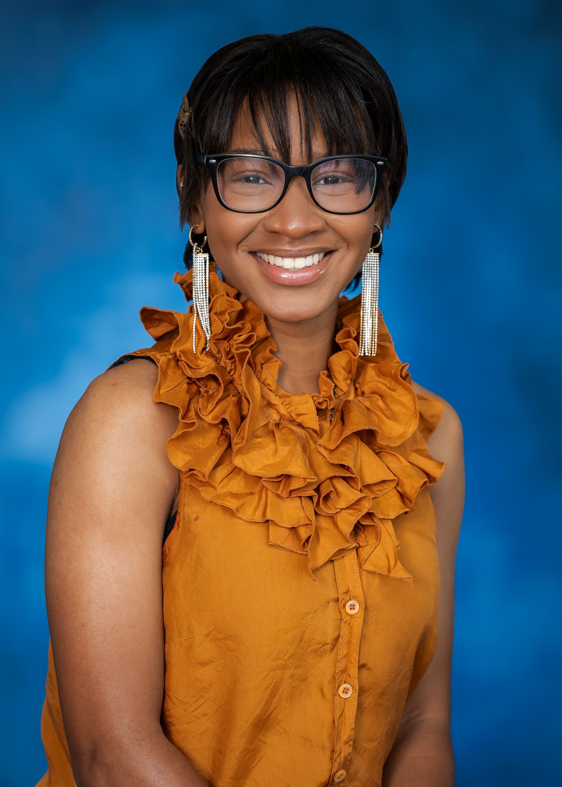 Woman with glasses, smiling, wearing an orange top with ruffled collar, against a blue backdrop.