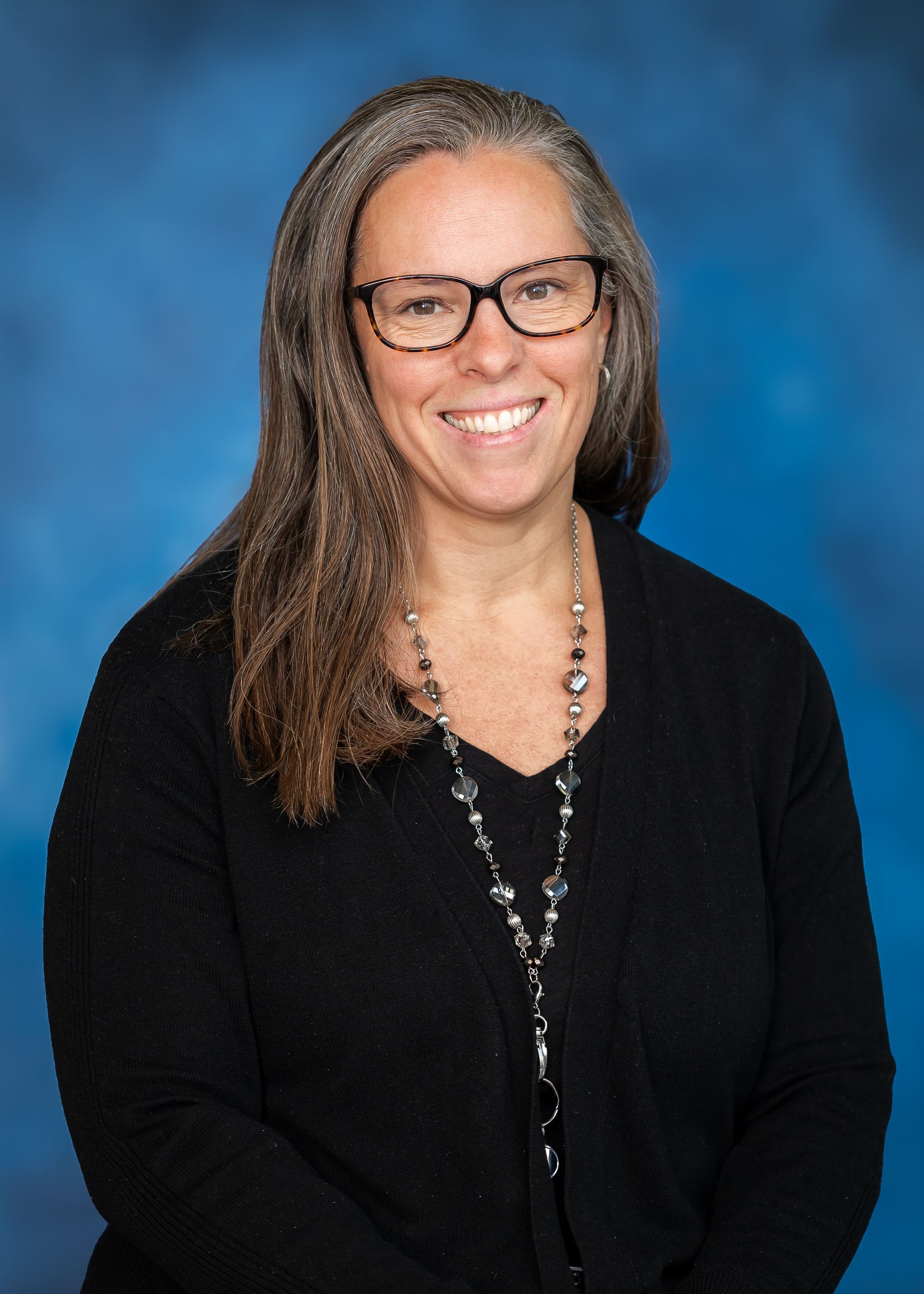 Woman with glasses, long gray hair, black sweater, and a necklace smiles against a blue background.