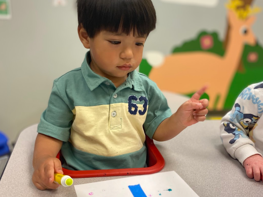Child in a red chair at a table, using a yellow marker to draw on white paper.