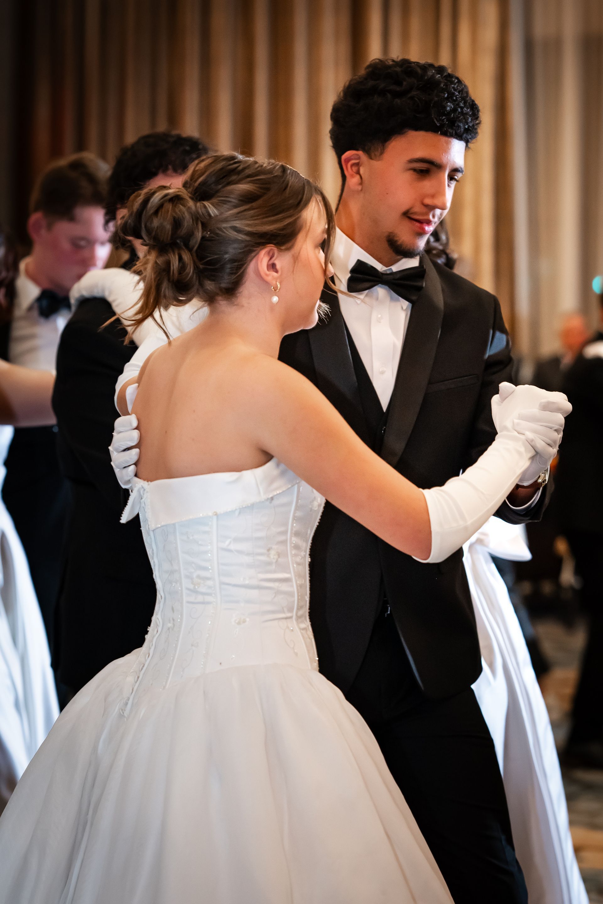 Couple in formal attire waltzes at a ballroom dance. They wear white gloves and the man a black tuxedo.