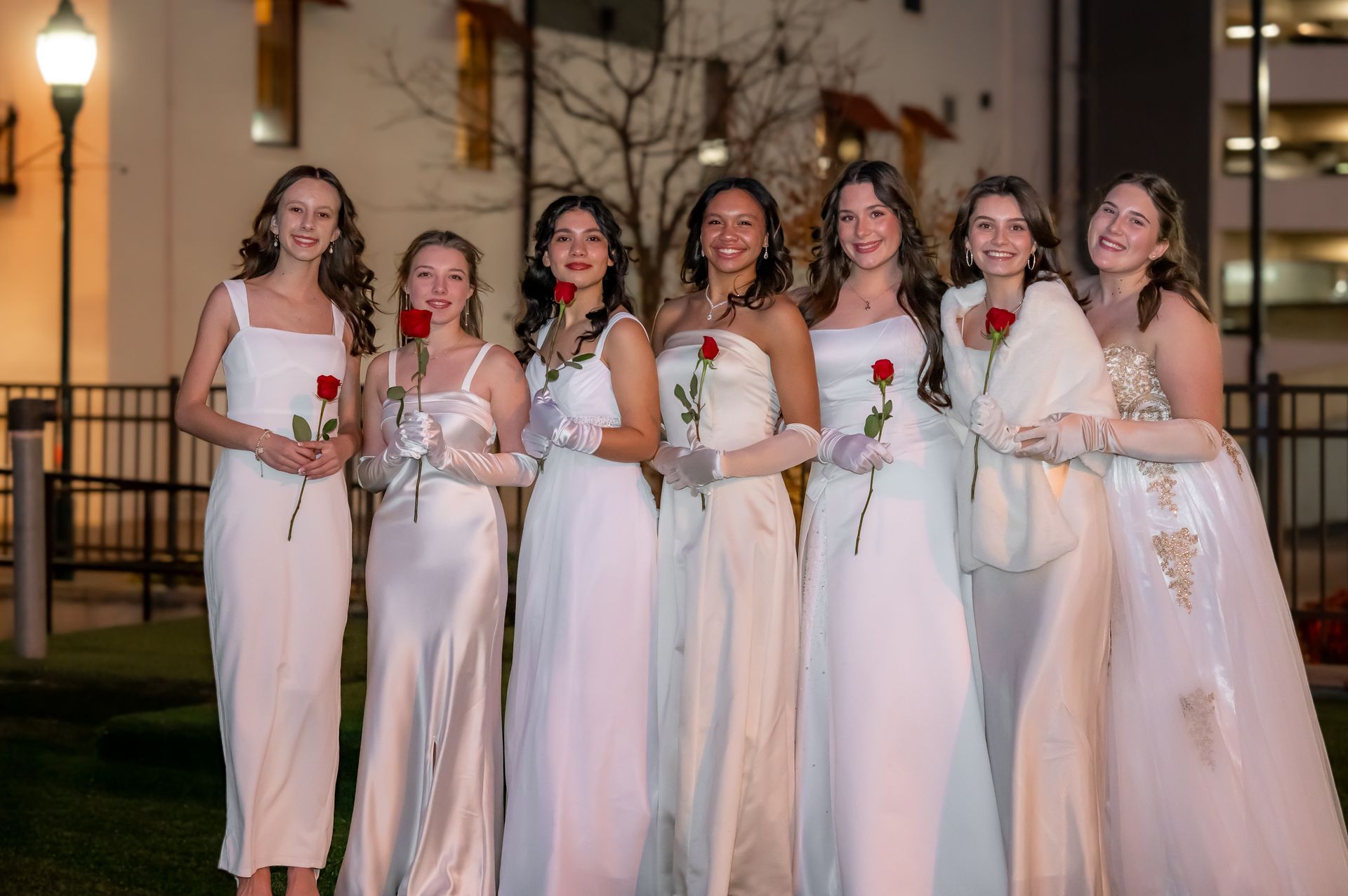 Seven women in white gowns holding roses pose outdoors at night.