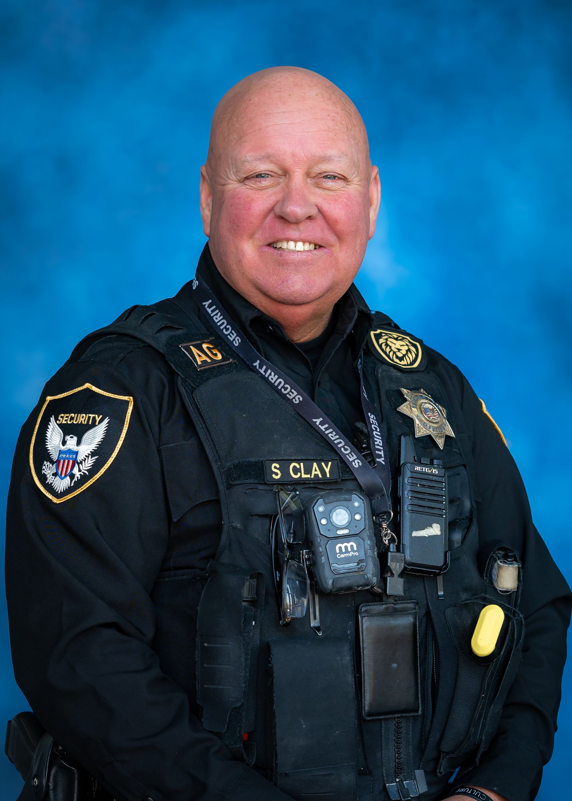 Man in security uniform; smiling. Wearing a badge, radio, and body camera.