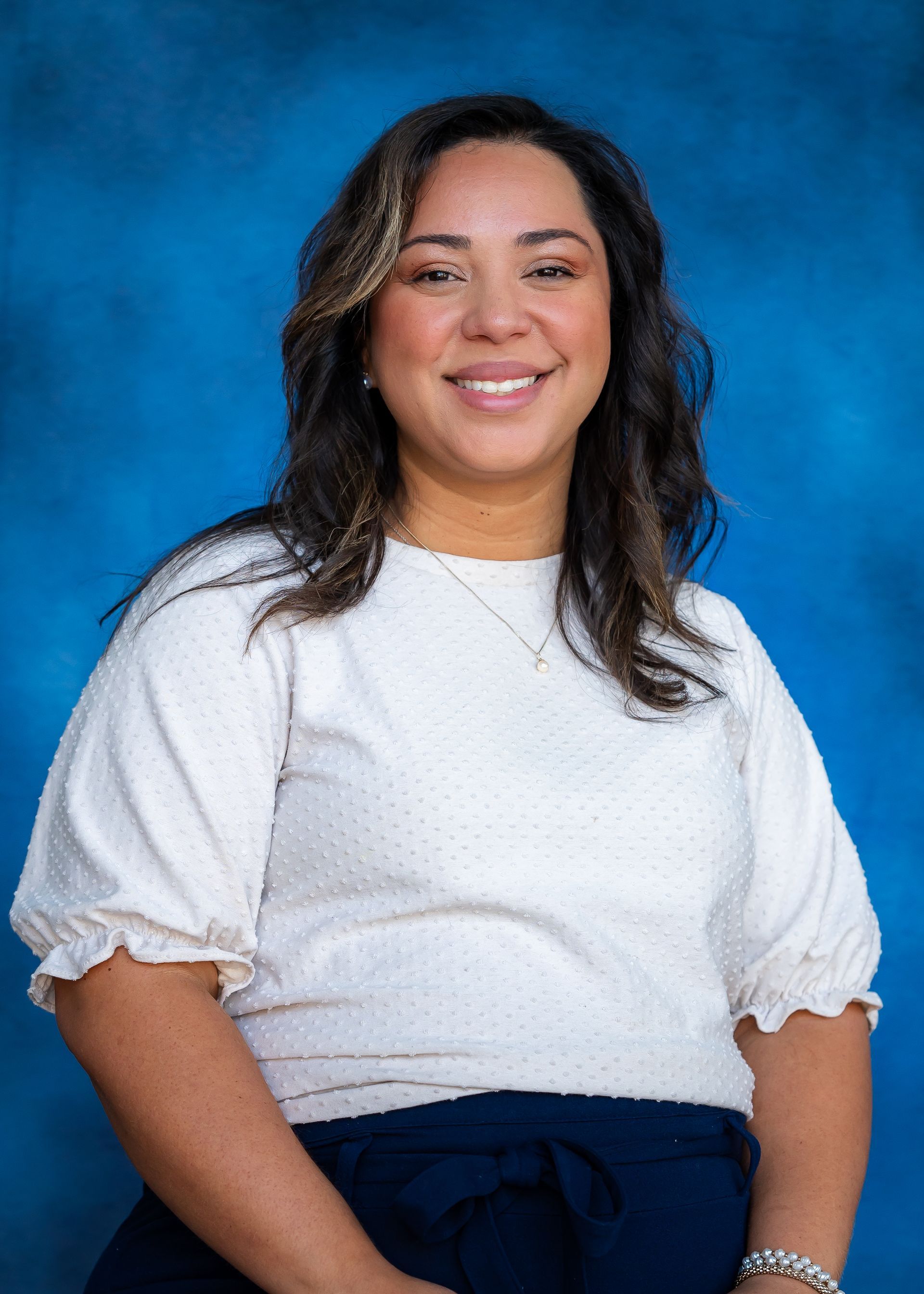 Woman with brown hair and a white shirt smiling against a blue background.