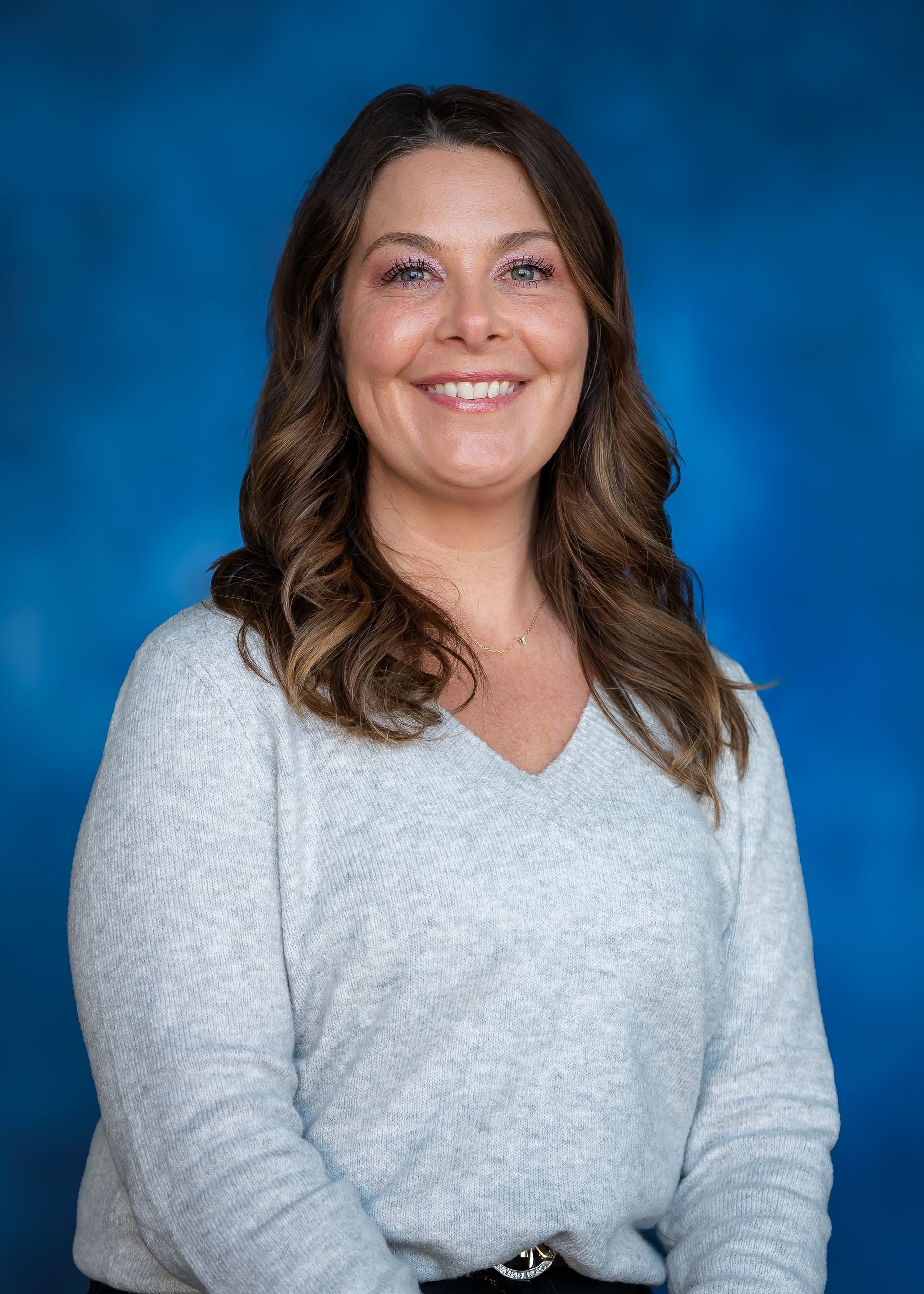 Woman with long wavy brown hair smiles at the camera, wearing a light gray sweater, in front of a blue background.