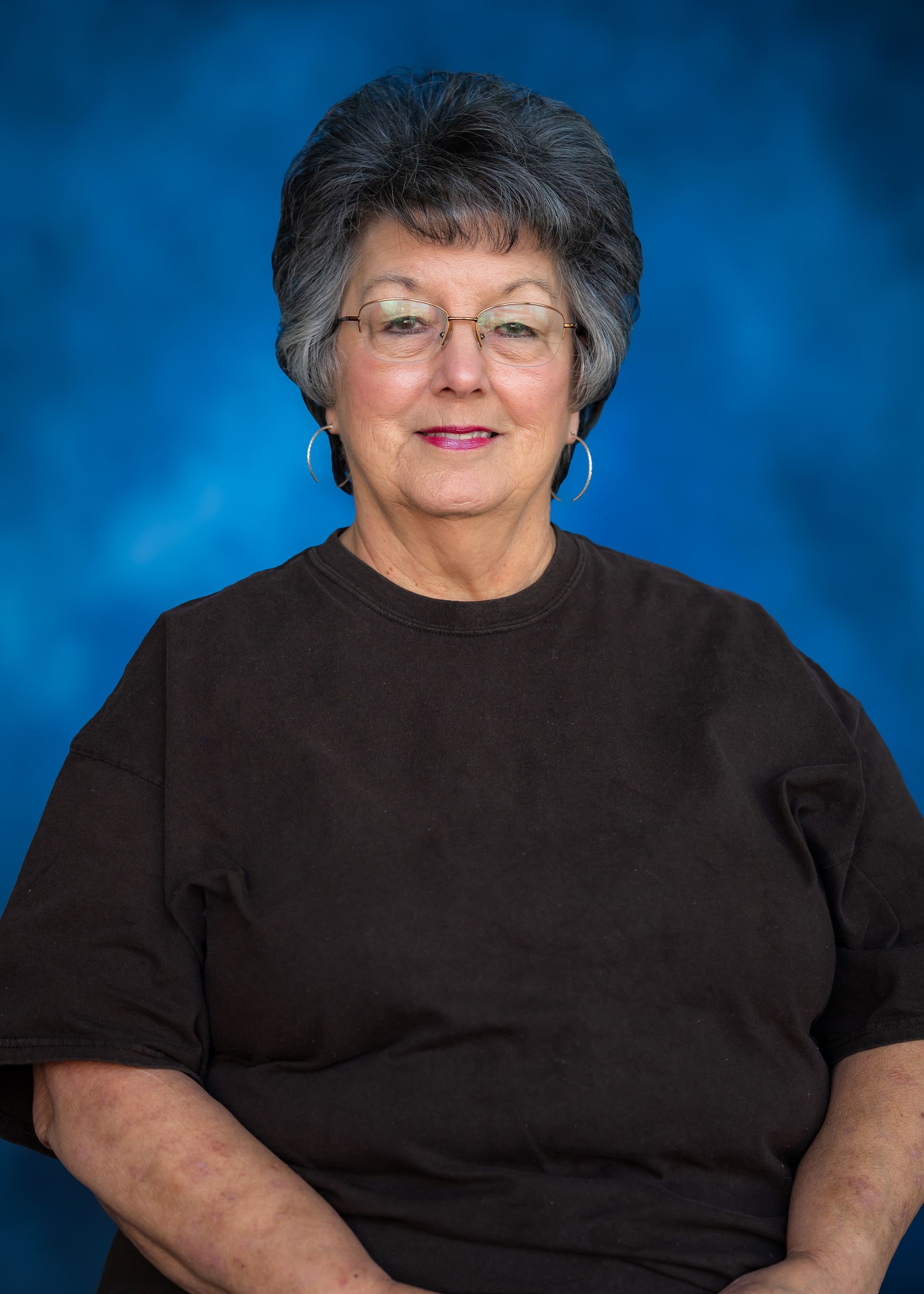 Woman with glasses smiling, wearing a dark brown shirt, against a blue background.