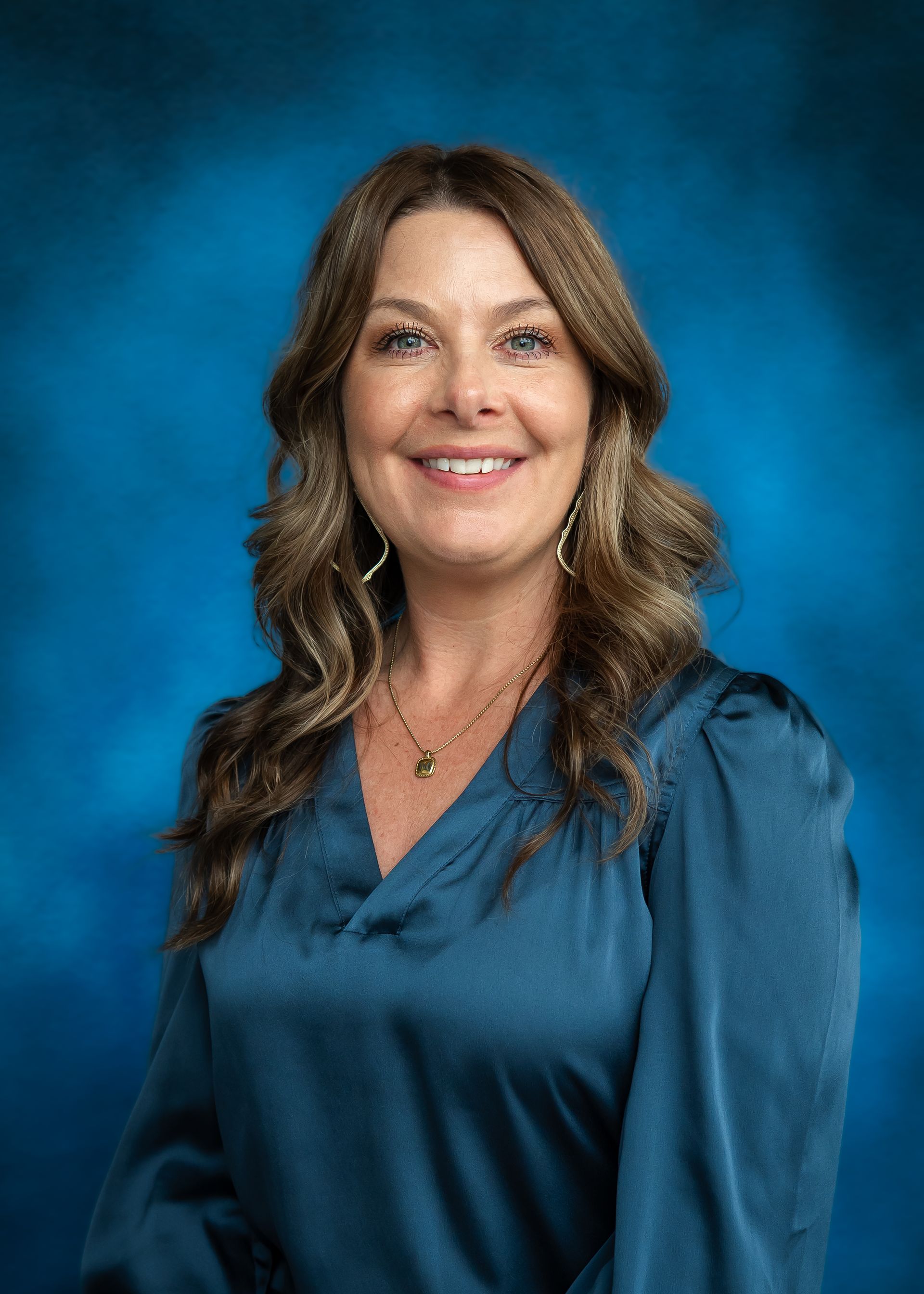 Woman with long wavy brown hair smiles at the camera, wearing a blue shirt, in front of a blue background.