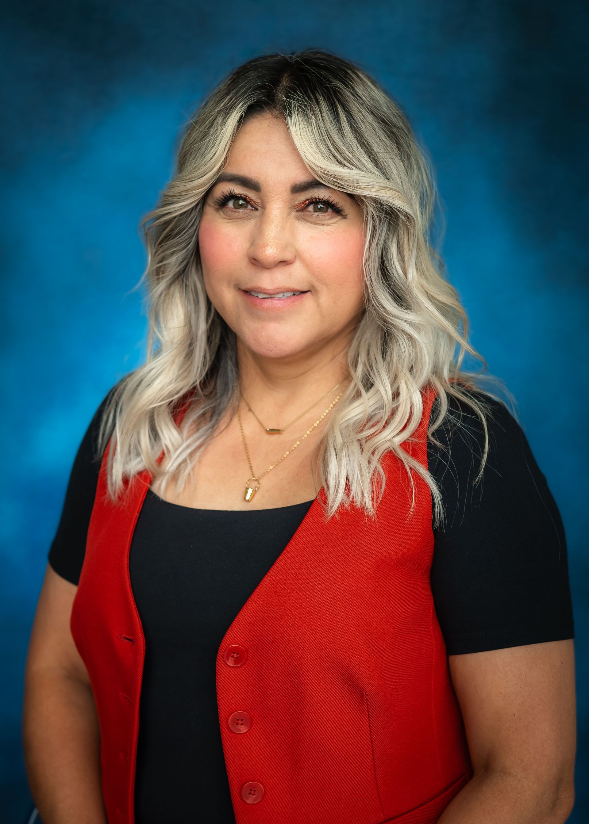 Woman with dark and blonde hair, wearing a black shirt with red vest, smiling, against a blue background.