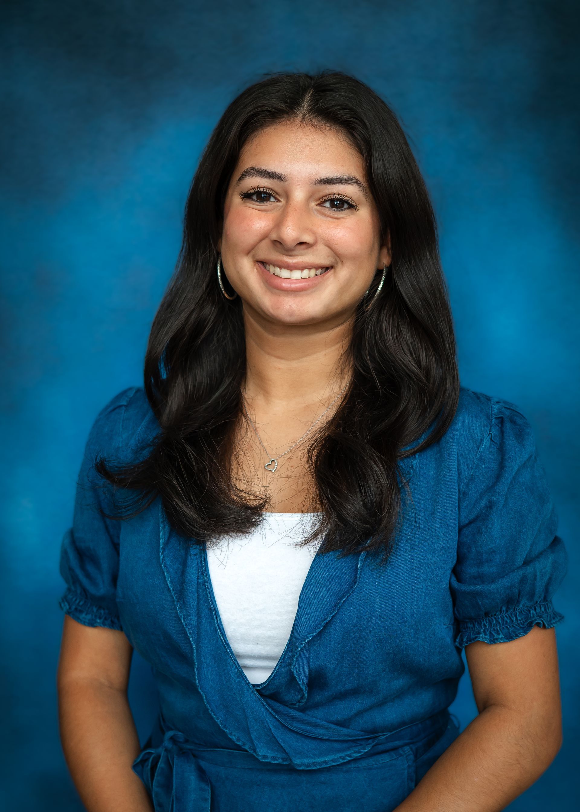 Smiling person with dark curly hair in a blue top with white underneath, against a blue backdrop.
