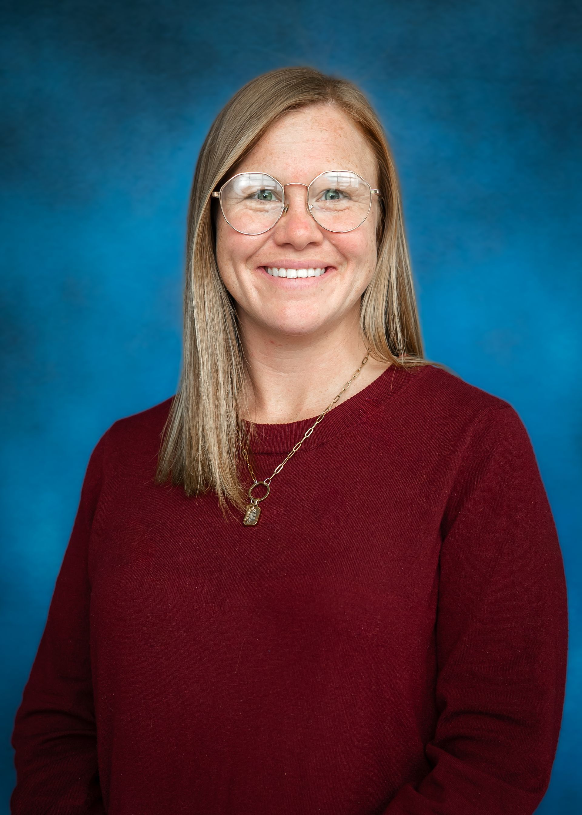 Woman with glasses, long auburn hair, wearing a maroon top, smiling against a blue backdrop.