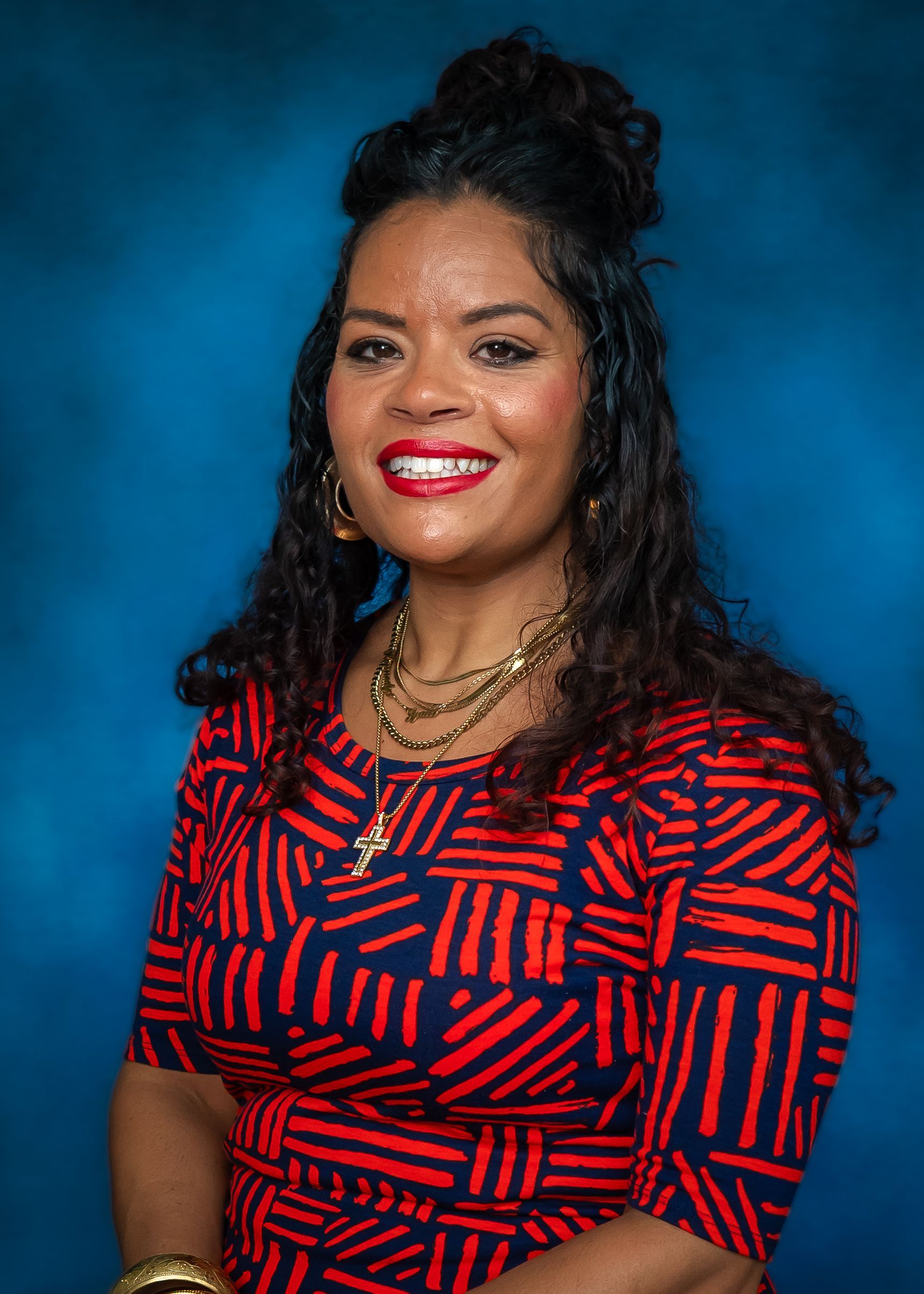 Woman with long curly hair, wearing a patterned blazer and smiling, against a blue backdrop.