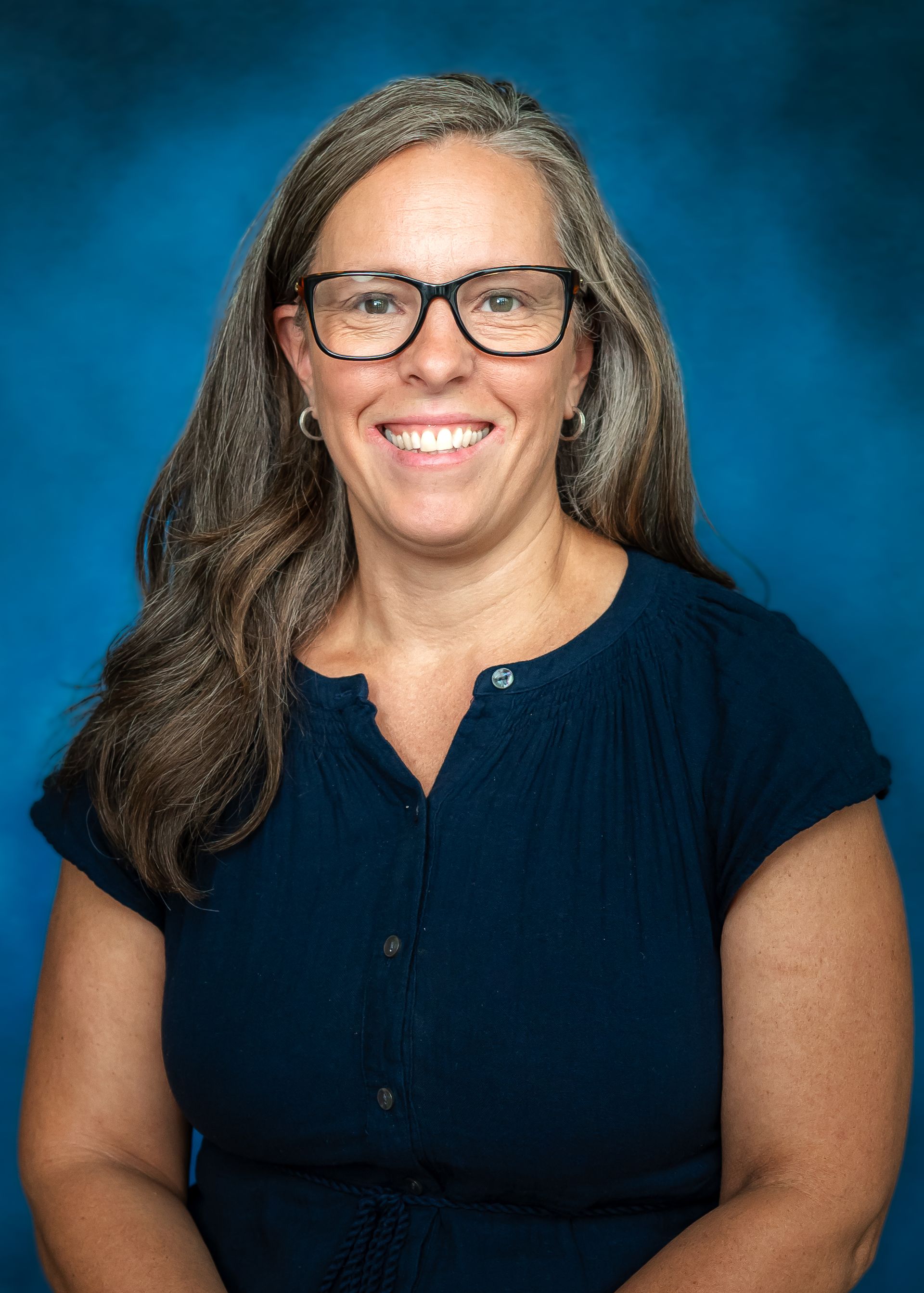 Woman with glasses, long gray hair, navy shirt, smiles against a blue background.