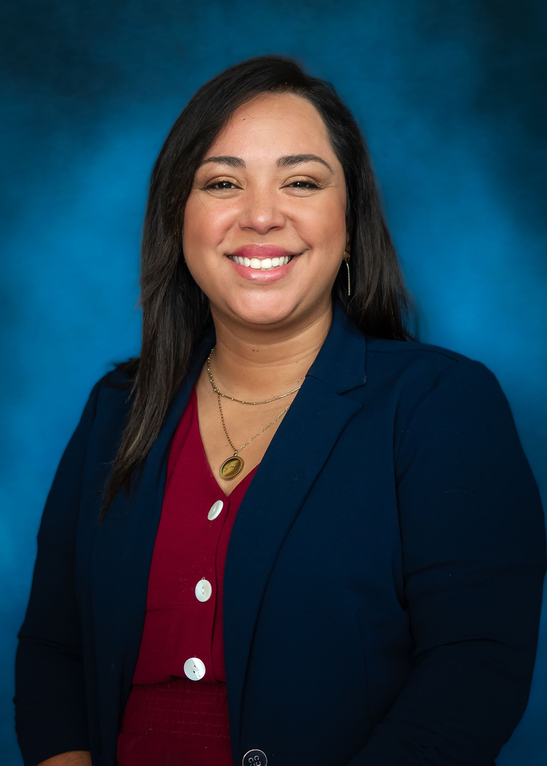 Woman with brown hair and a navy jacket and maroom shirt smiling against a blue background.