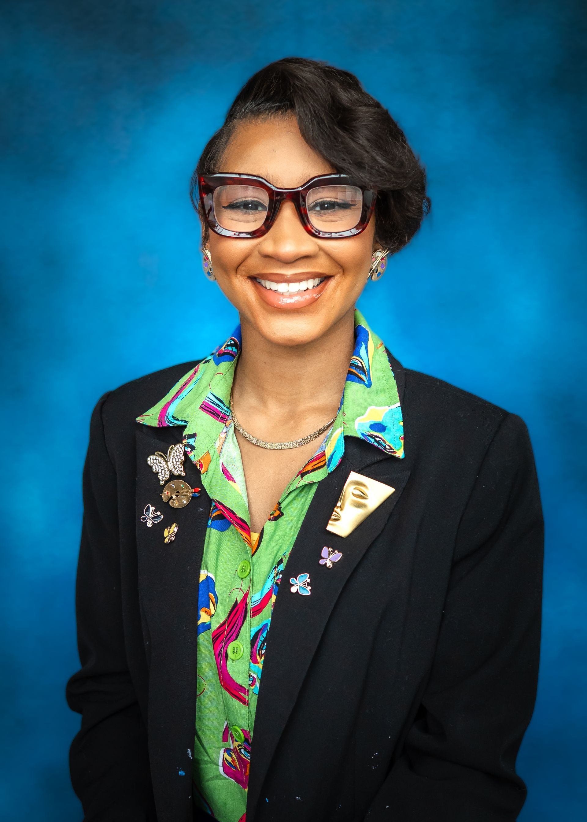 Woman with glasses, smiling, wearing a navy blazer, green and colorful print blouse with collars, against a blue backdrop.