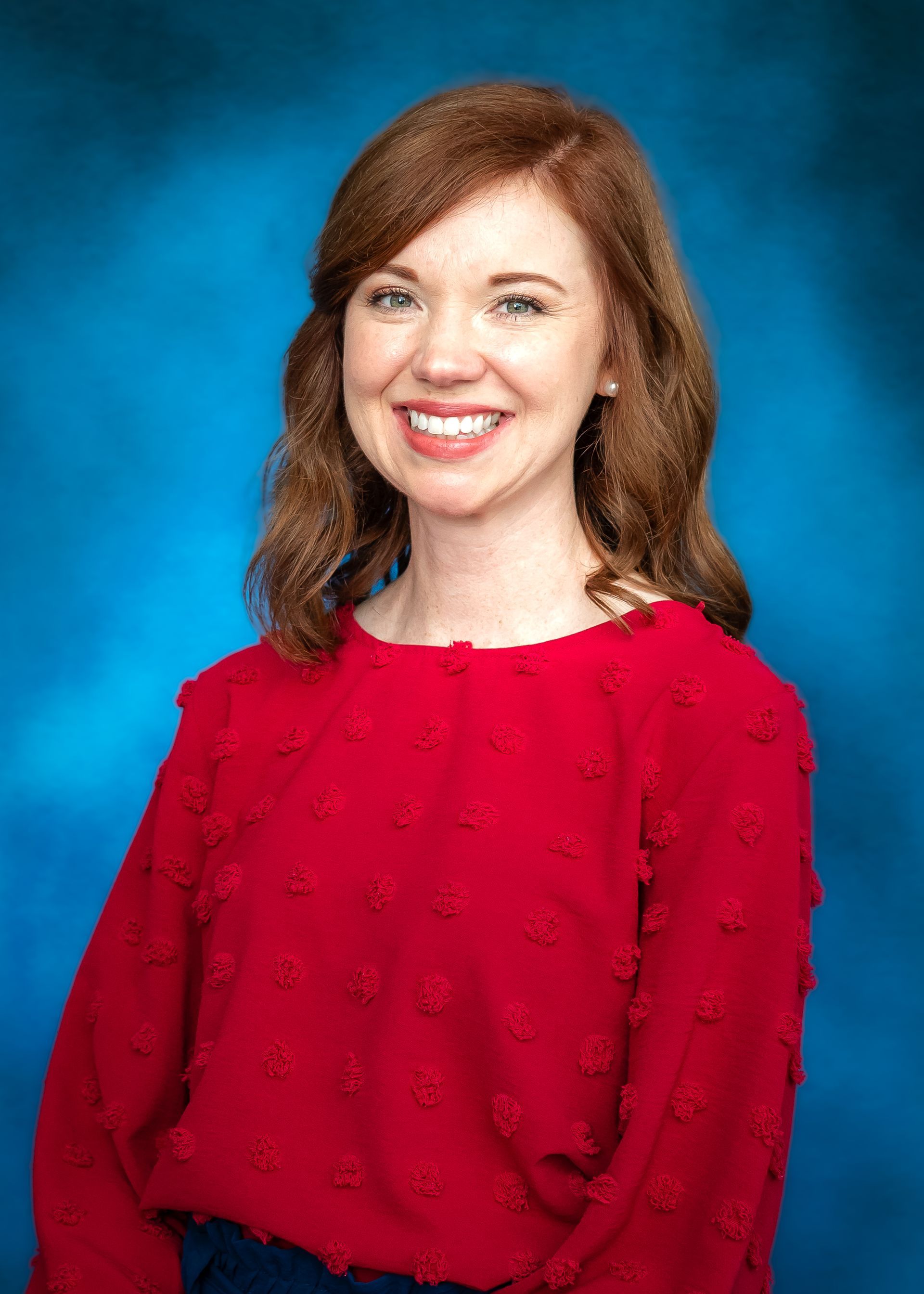Woman with red hair smiling, wearing a red shirt against a blue background.