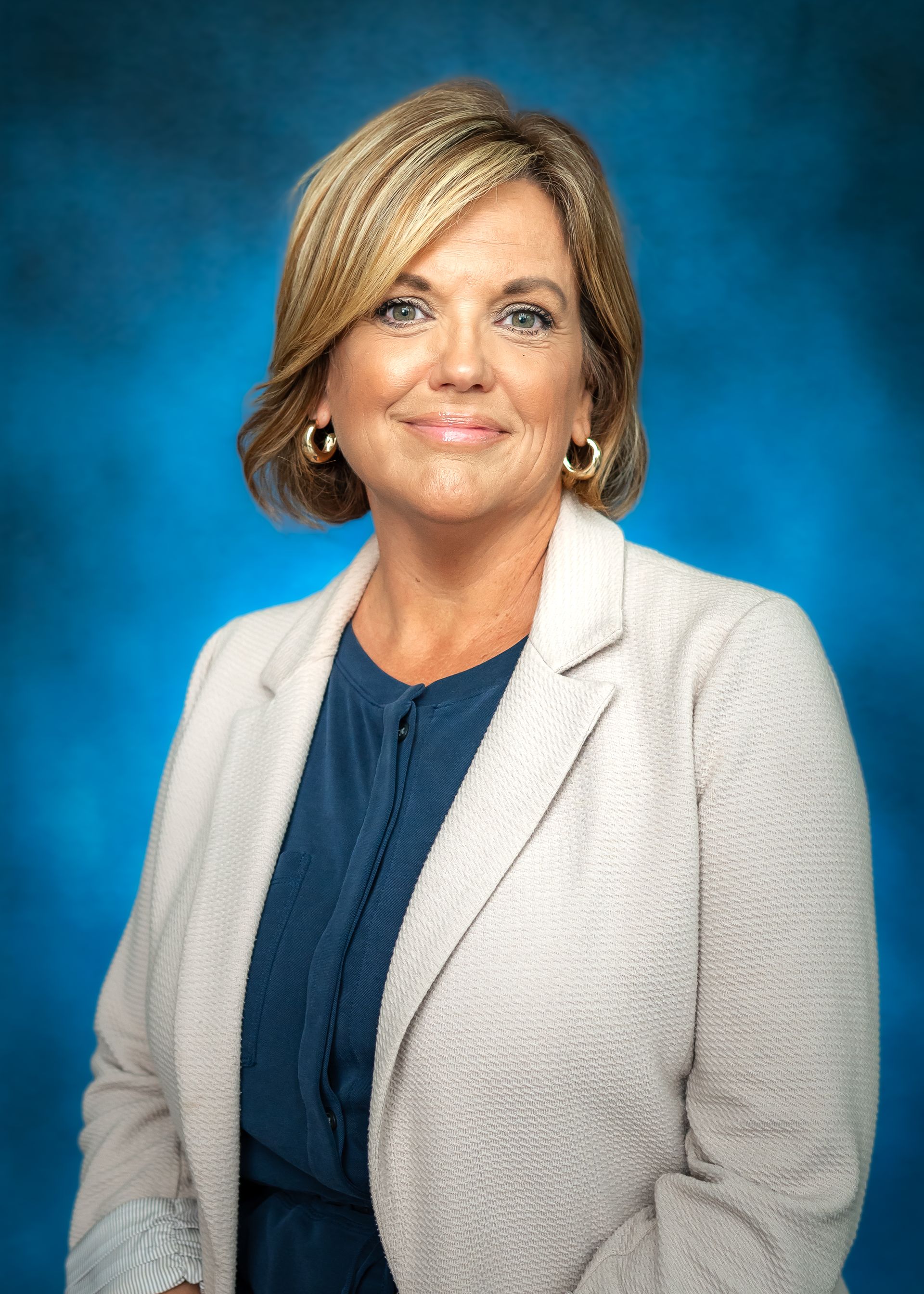 Woman with short brown and blonde hair smiles, wearing a cream jacket with a dark blue top, against a blue background.
