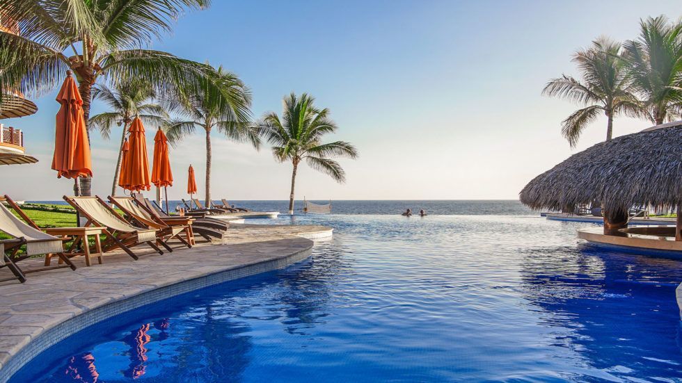 Vivo Resort poolside scene: blue pool, ocean view, palm trees, orange umbrellas, lounge chairs.