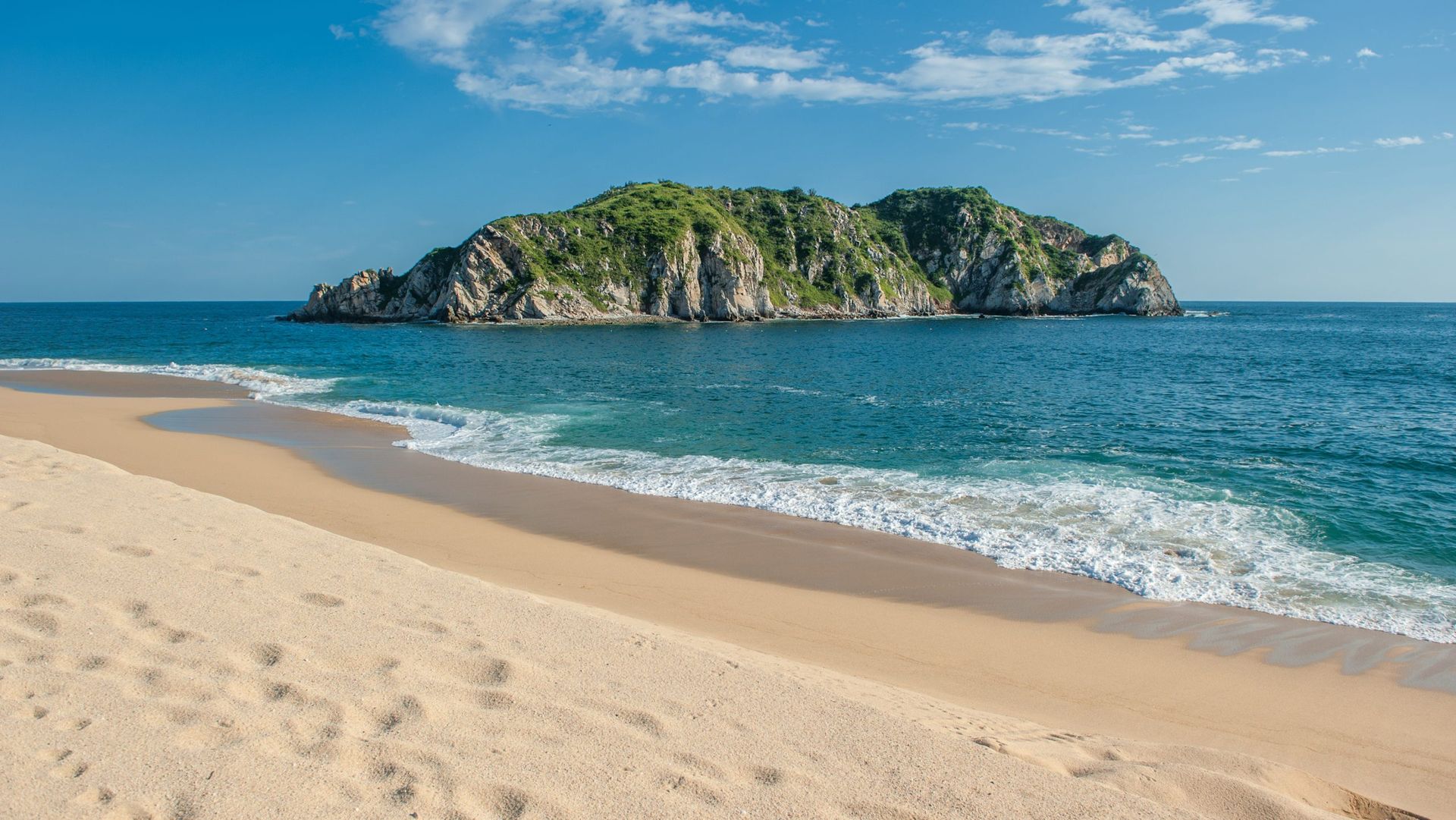 Sandy beach with small island and blue ocean under clear sky.