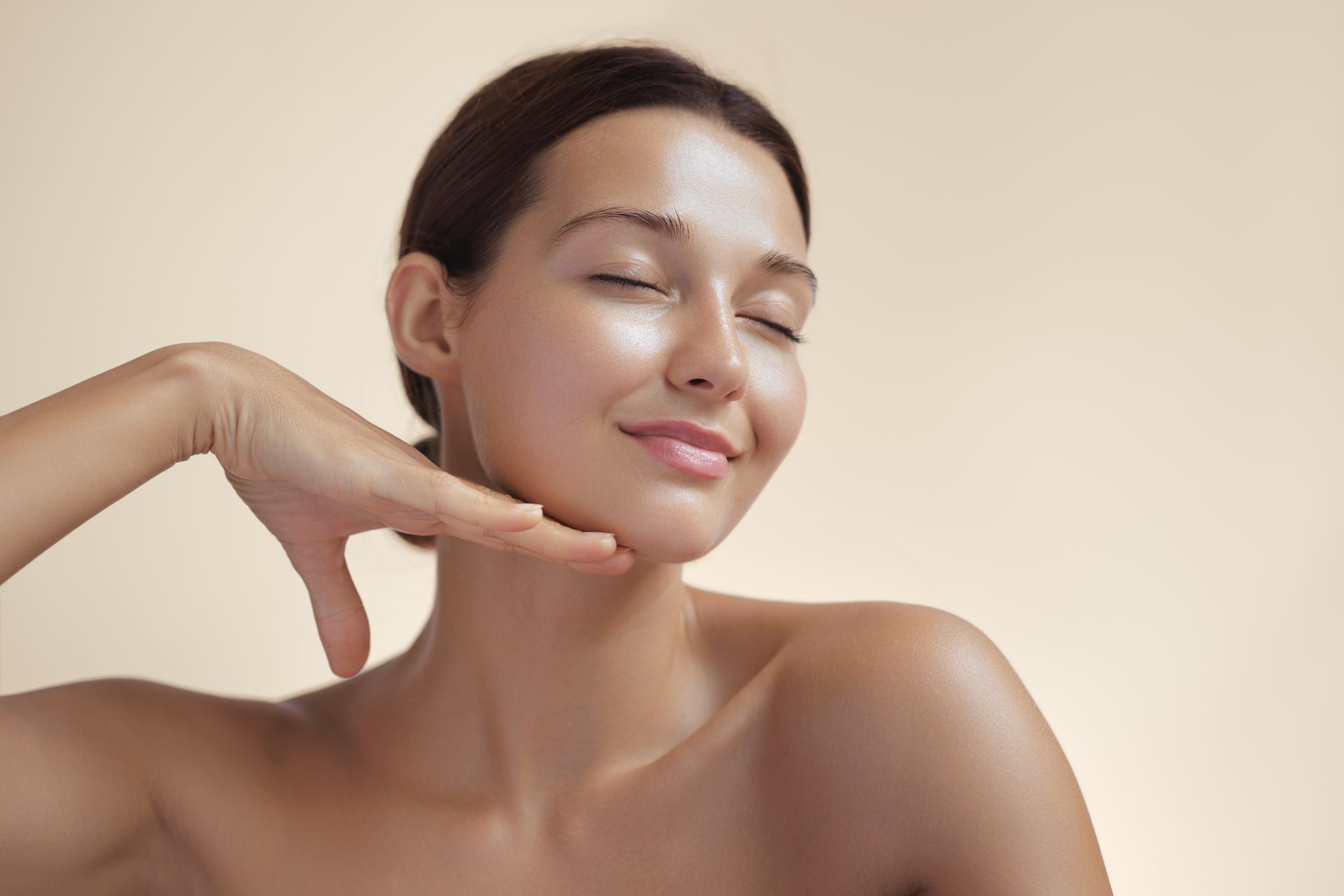 Woman with glowing skin, hand on jawline, eyes closed, smiling, beige background.