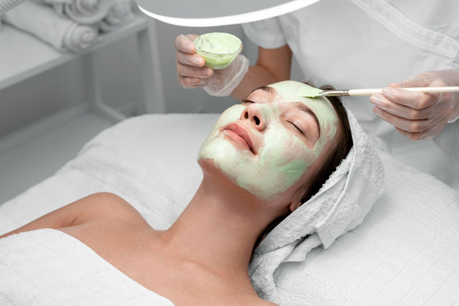 Woman getting a facial mask applied at a spa; hands in gloves, green mask, bright light.