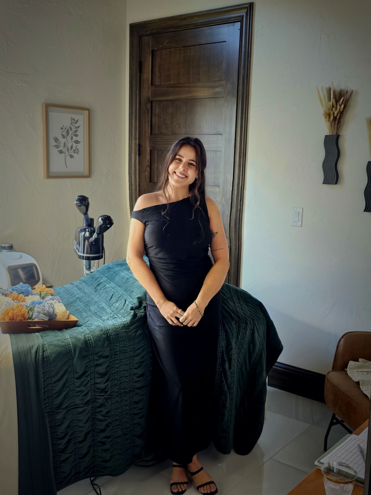 A woman in a black dress is standing next to a bed in a room.