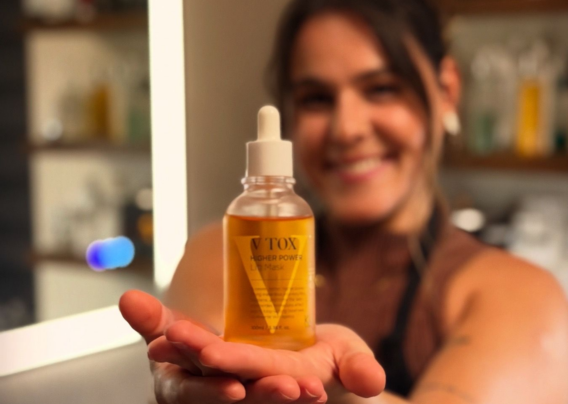 Woman smiling, holding a skincare oil bottle, blurred background of a bathroom.