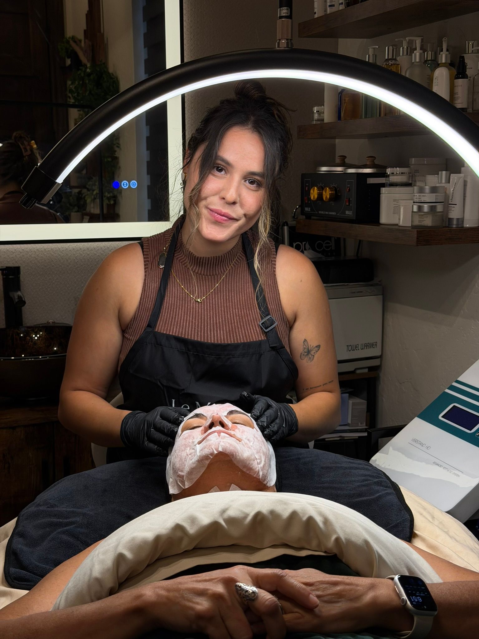 Woman giving facial treatment. Both wear black gloves. Overhead light illuminates room.