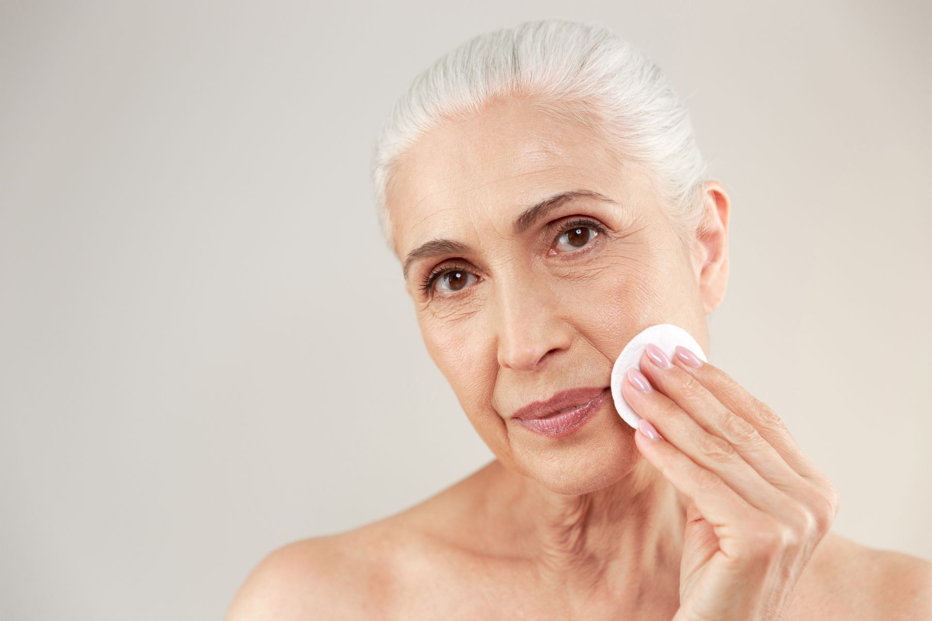 Woman with gray hair, using a cotton pad to cleanse her cheek against a plain background.