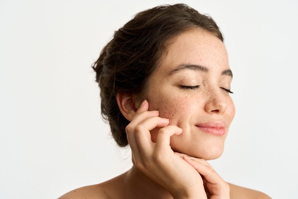 Woman with freckles, eyes closed, smiling, touching her face, white background.