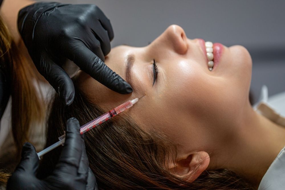 Woman receiving a facial injection, smiling. A hand in a black glove holds the syringe near her eye.